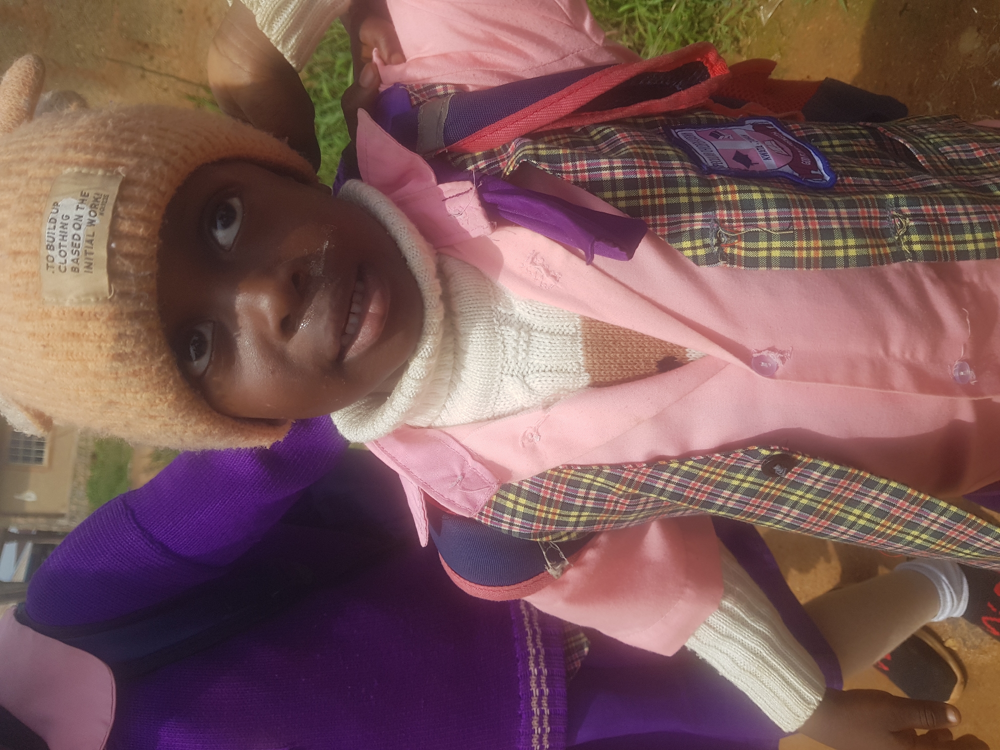 A medium shot shows a young Black child wearing a tan knitted hat with ear flaps, a cream-colored turtleneck sweater, and a pink school uniform jacket. The jacket has a plaid vest overlay. A backpack with red and purple straps is visible. The child is looking at the camera with a slight smile. The background is out of focus, showing a building and green grass. The scene appears to be outdoors in a rural or suburban setting.