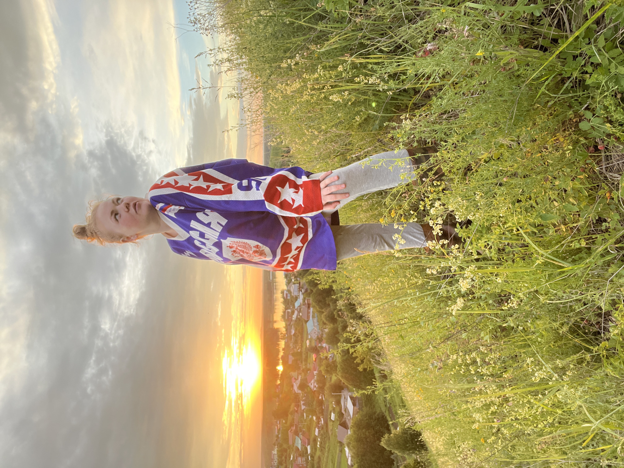 A young Caucasian woman with blonde hair tied in a bun stands on a grassy hillside overlooking a village at sunset. She is wearing a blue and white hockey jersey with red accents and stars, gray leggings, and brown boots. The sun is setting behind a body of water, casting a warm glow over the village. The sky is filled with clouds. The hillside is covered in green grass and wildflowers. The village appears to be in a rural area of Russia.
