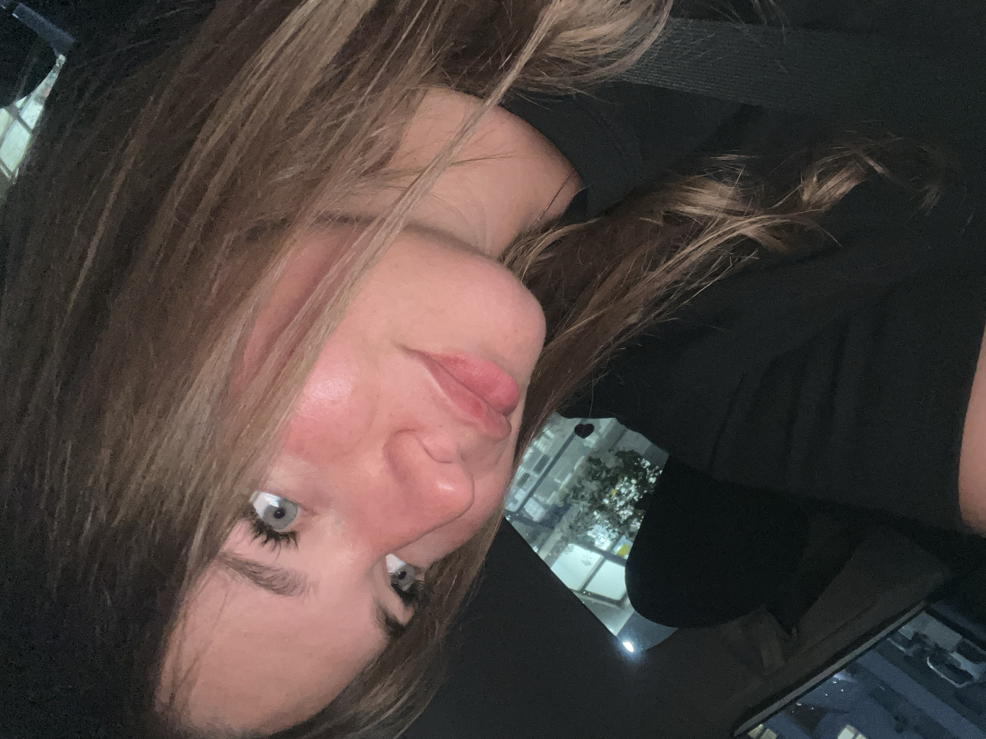 A close-up, tilted portrait features a young woman inside a vehicle, likely a car, during the evening or night. Her face is positioned diagonally across the frame, with her light blue or grey eyes looking directly at the viewer. She has light brown hair, which cascades around her face and extends into the foreground and background. Her lips are slightly parted.
She is wearing a dark-colored top or hoodie, parts of which are visible. The setting is confirmed as inside a car by the dark interior elements and reflections visible. Through a window or in a reflection on the right side, an urban environment can be seen, characterized by illuminated buildings and what appears to be trees or foliage. A small, dark heart-shaped object hangs in this reflection. The ambient lighting is dim, suggesting it's nighttime or late evening, with artificial lights illuminating the exterior scene. Her expression is calm and direct. No text is visible in the image. The location context for the scene is Budva, Montenegro.