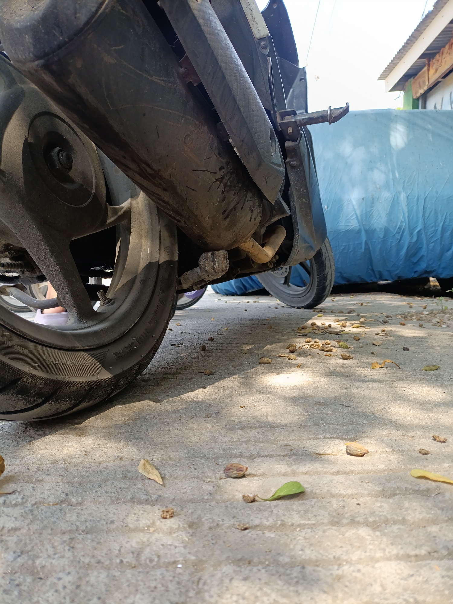 A low-angle, close-up shot captures the rear portion of a motorcycle, focusing on the exhaust pipe and wheel. The motorcycle is parked on a paved surface, possibly a street or driveway, in Cariu, Indonesia. Scattered dry leaves and small debris are visible on the ground, suggesting an outdoor setting. In the background, a large blue tarp covers an unseen object, and a portion of a building with a corrugated metal roof and decorative elements is visible. The lighting indicates it is daytime, with distinct shadows cast by the motorcycle and other objects. No people are visible in the frame, and no specific actions are depicted, other than the motorcycle being stationary. The overall impression is one of an ordinary outdoor scene in a residential or commercial area of Cariu.
