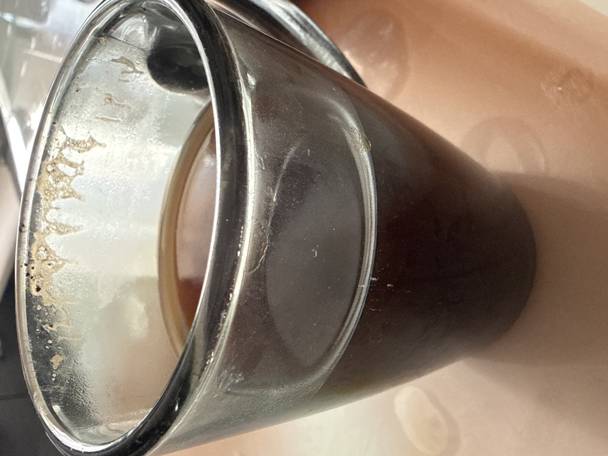 The image shows a close-up of a clear glass mug filled with dark liquid, likely coffee. The mug has some residue around the rim and on the interior walls. The mug is placed on a light-colored, possibly ceramic surface, which has a subtle texture and appears to be a tabletop. The background is blurred, suggesting shallow depth of field. There are no people or animals visible in the image. The lighting appears to be natural, casting soft shadows. No specific location in Simferopol, Ukraine, can be determined from the visual information.