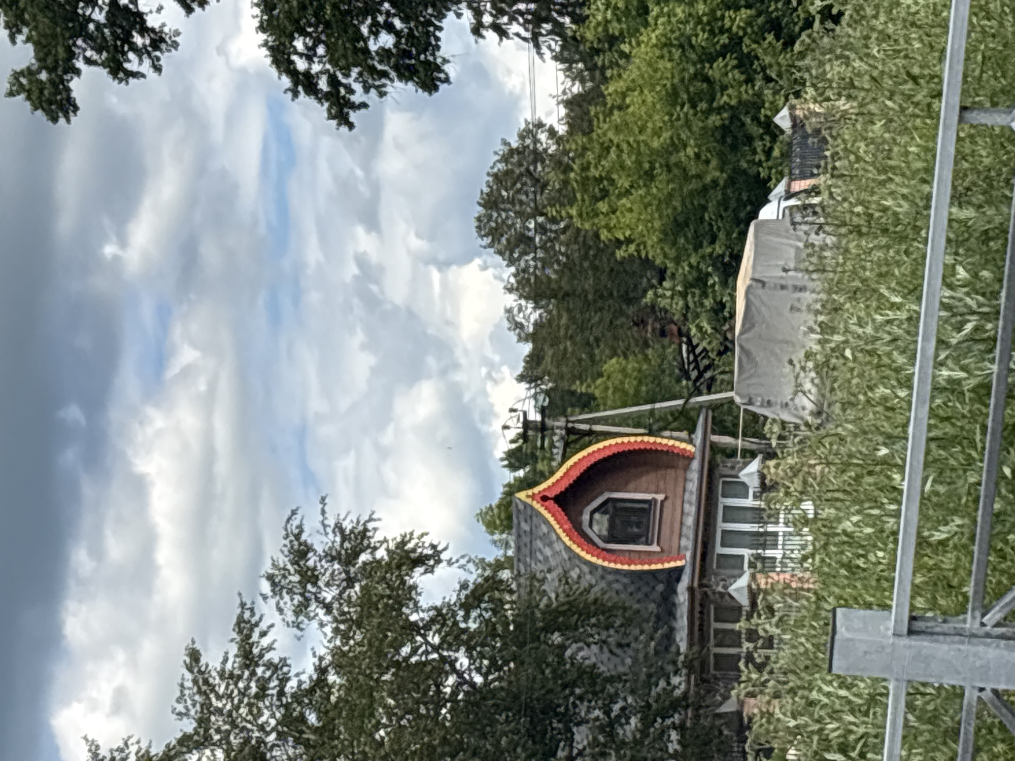 The media file depicts a cloudy sky with dramatic, overcast conditions. Dark grey clouds fill a significant portion of the frame, interspersed with patches of lighter grey and hints of pale blue sky. The tops of lush green trees frame the upper edges of the image, their leaves appearing dense and full.
In the mid-ground, a distinctive building with a unique roofline is visible. The roof is steeply pitched and adorned with decorative red and yellow trim, giving it a whimsical or traditional architectural style. A small, dark window is set into the roof's dormer. The building itself appears to be constructed with grey shingles or siding. Behind this main structure, a portion of another building with white-framed windows can be seen, suggesting a residential or perhaps a guesthouse setting. Part of a large structure covered in a grey tarp is also visible, hinting at ongoing construction or perhaps protection from the elements.
To the right of the frame, a metal structure, possibly part of a fence or scaffolding, runs vertically. Dense green foliage, likely more trees or bushes, extends along the right side and partially obscures the buildings. The overall impression is one of a slightly overcast day in a semi-rural or suburban setting, with the unique building's architecture being a focal point. The time of day is difficult to ascertain due to the overcast sky, but the lighting suggests it is daytime. There is no visible text in the image.
