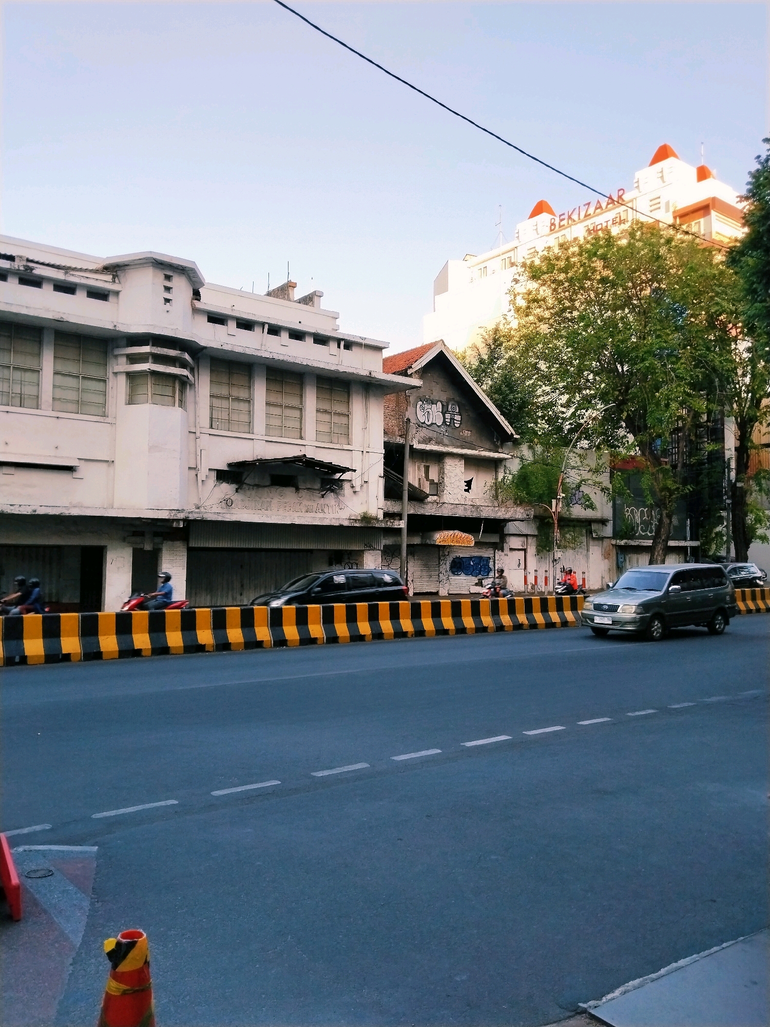 The image depicts a street scene in Surabaya, Indonesia, under a clear sky. In the foreground, a gray minivan is driving on an asphalt road. A yellow and black striped barrier runs along the side of the road, separating it from the sidewalk area. Behind the barrier, a building with a white facade and large windows is visible. Further down the street, another building with a weathered facade and graffiti can be seen. The word "BEKIZAAR" is prominently displayed in red letters on the upper portion of a white building in the background, which also has red-capped roofs. Several trees with green foliage are present, some partially obscuring the buildings. Two people are riding on a motorcycle in the middle ground, and another person is on a red motorcycle further back. The scene appears to be captured during the day, possibly in the late afternoon given the lighting.
