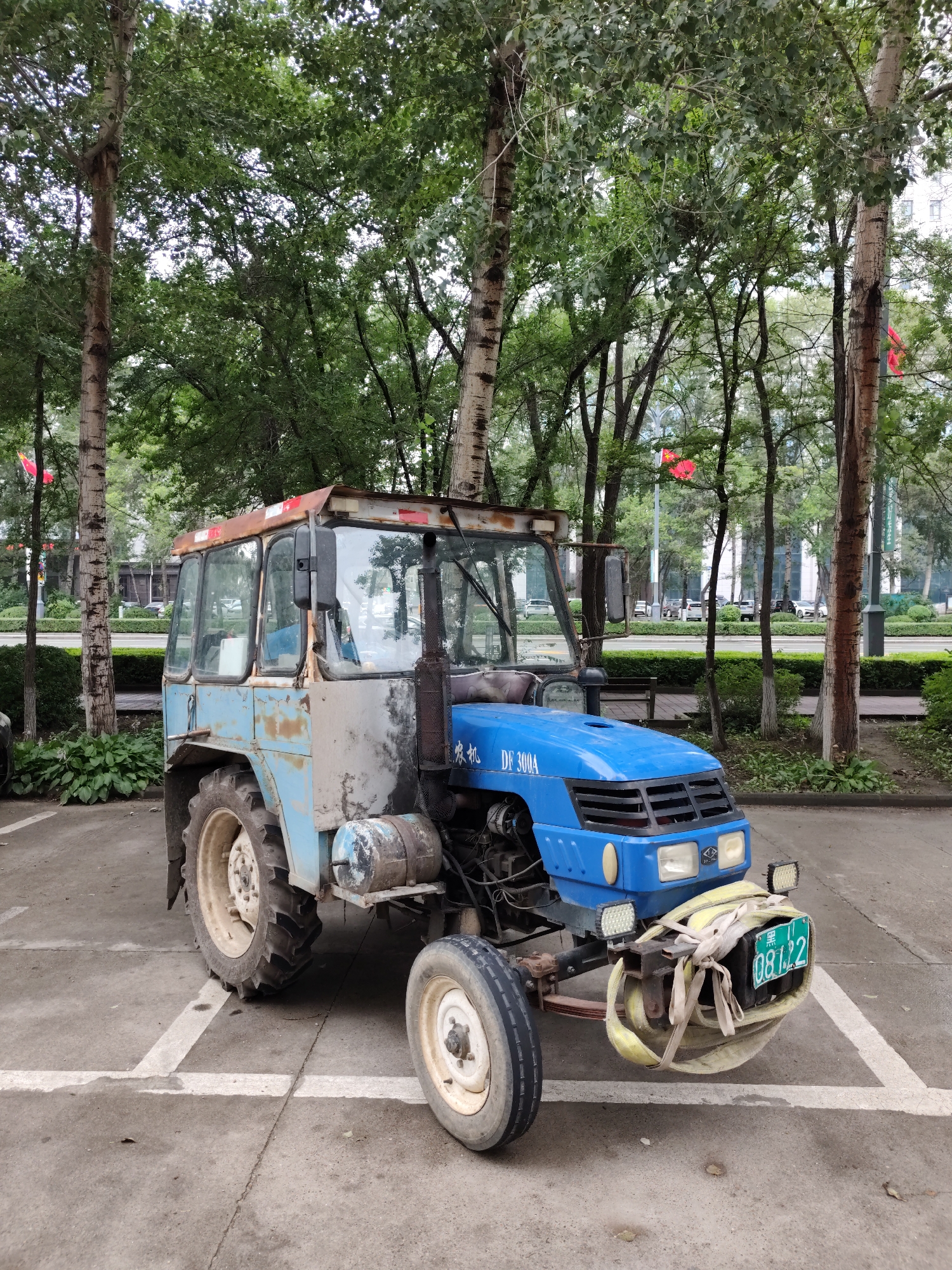 A blue and weathered light blue tractor is parked on a concrete surface marked with white lines in 幸福乡, China. The tractor has a cab with multiple windows and a rusty metal frame. The front of the tractor is blue and has the model name "DF 300A" inscribed on it. A license plate with Chinese characters and the number "08 L22" is attached to the front bumper, which is fitted with yellow straps. The tractor has large rear wheels and smaller front wheels. It is surrounded by trees with green foliage, and in the background, there are more trees, some buildings, and a road with parked cars. The ground appears to be somewhat wet in places.