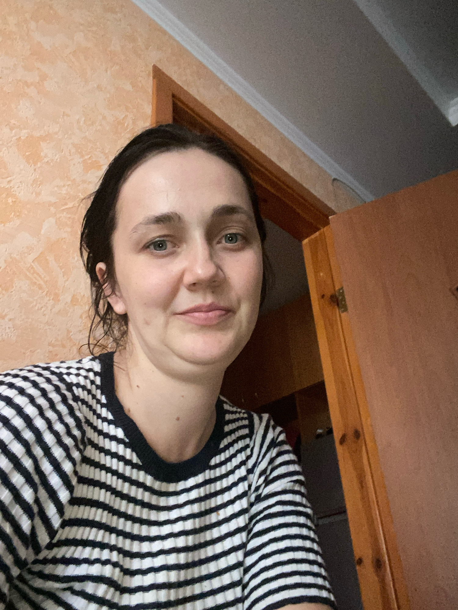 A young woman with dark hair and light blue eyes is photographed from the chest up, indoors. She is looking directly at the camera with a slight smile. She is wearing a black and white striped shirt. Behind her, a portion of an orange textured wall and a wooden door frame are visible, as well as a sliver of a grey ceiling. The scene suggests a domestic interior.