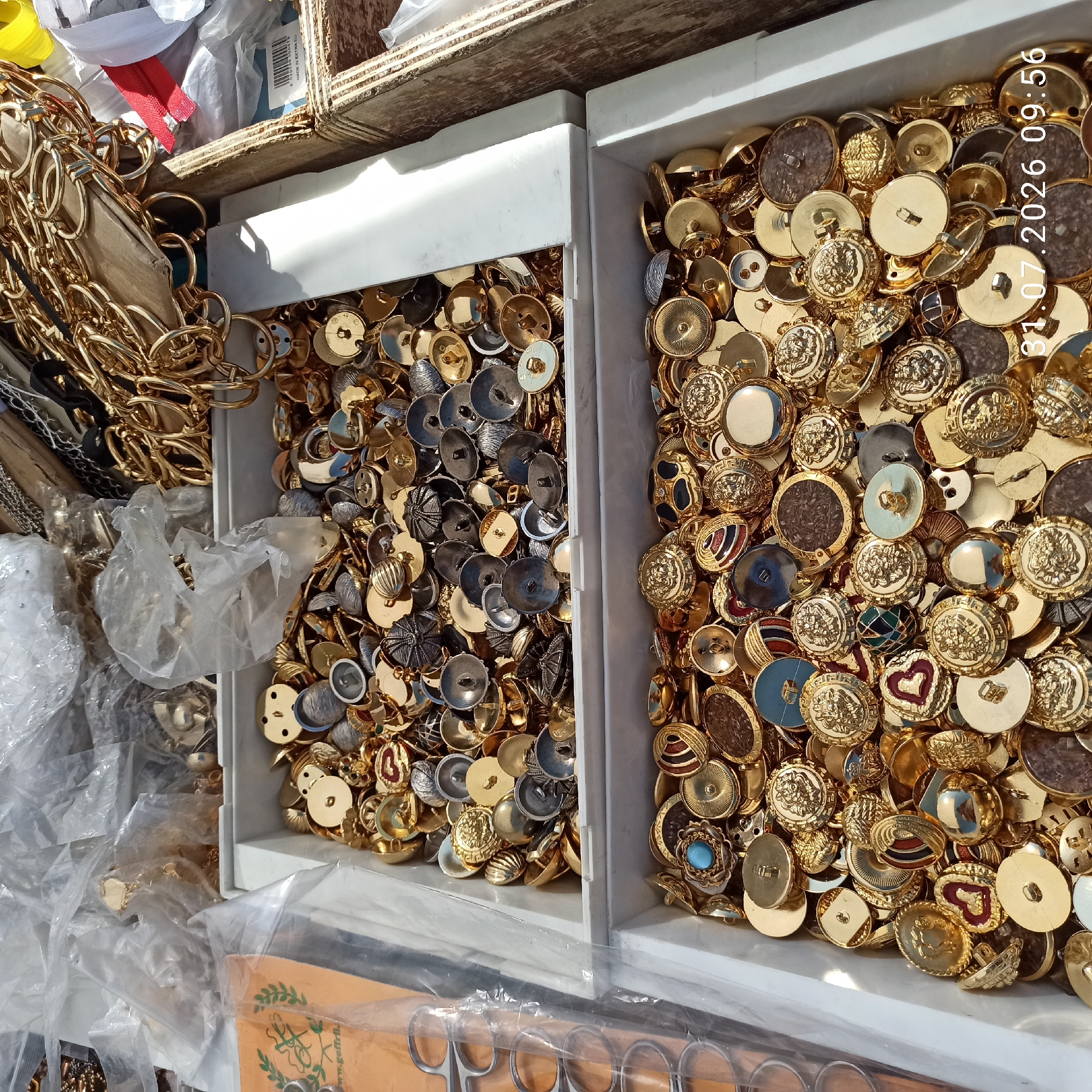 The image depicts a close-up, overhead view of two white plastic bins filled with an assortment of golden buttons and other hardware. The buttons vary in size, design, and material, with many featuring intricate patterns, some with enameled details, and others appearing to be plain metal. There are also some grey buttons visible. In the background, out of focus, are clear plastic bags containing more items, as well as a large quantity of gold-colored chain links and a red zipper. The lighting suggests an outdoor or well-lit indoor setting. The timestamp "31.07.2026 09:56" is superimposed on the lower right portion of the image. There are no people visible. The context suggests a market stall or a craft supply area in Modugno, Italy.