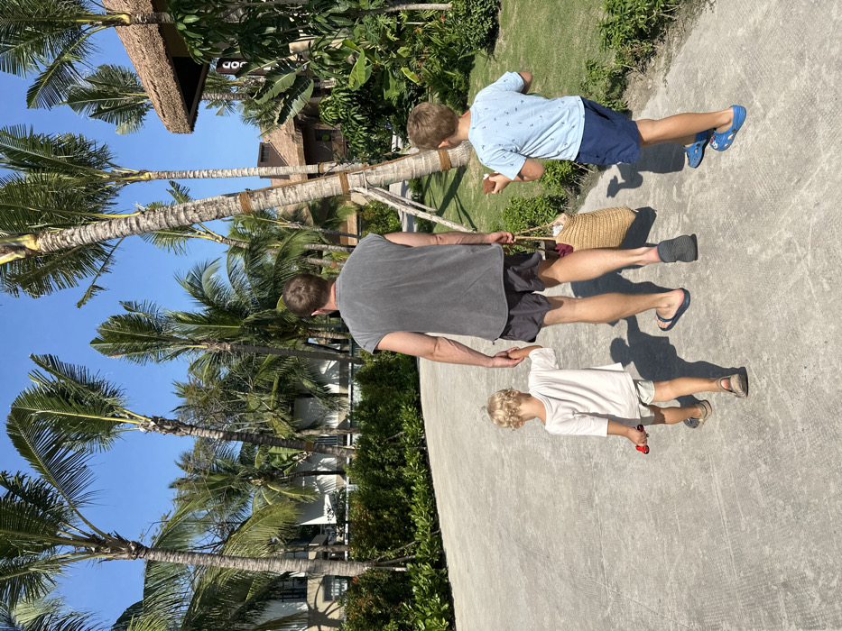 Three individuals, seen from behind, walk along a light grey paved pathway under a clear blue sky in a resort area of Phú Quốc, Vietnam.
The central figure is an adult male, wearing a faded dark grey sleeveless t-shirt, dark grey shorts, a blue flip-flop on his left foot, and a dark grey slide on his right. He carries a woven light brown bag in his right hand.
To his immediate left, a young boy with blonde curly hair, wearing a light-colored t-shirt and shorts with light brown sandals, holds the adult's left hand. He holds a small red toy in his right hand.
To the adult's right, an older boy with short brown hair, wearing a light blue patterned t-shirt, dark blue shorts, and blue Crocs, walks with a piece of coconut shell in his right hand.
The pathway curves slightly right, bordered by green, manicured vegetation. To the left, several multi-story buildings with light facades and dark roofs are visible behind hedges. To the right, resort-style buildings with thatched roofs are discernible amidst dense tropical foliage and numerous tall palm trees. One building on the right has a partial sign showing "CO". Sunlight illuminates the scene, casting shadows on the pathway.