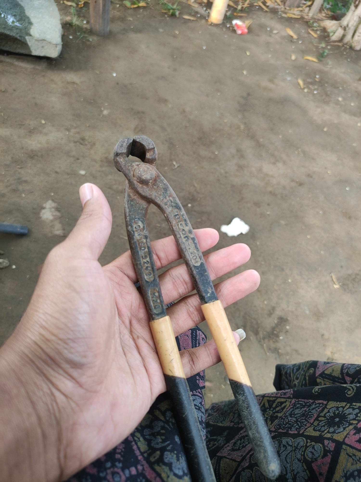 An outdoor, eye-level, close-up shot shows a pair of rusty bolt cutters held in a person's left hand. The bolt cutters are old and show signs of wear and tear, with a light-colored grip and black handles. The person's hand is slightly cupped around the handles. In the background, the ground is a mixture of dirt and scattered leaves, with a large rock on the left and some blurry foliage and wooden posts visible on the right. The overall scene appears to be in a rustic or rural setting, potentially in the city of Tiudan, Indonesia, given the context.