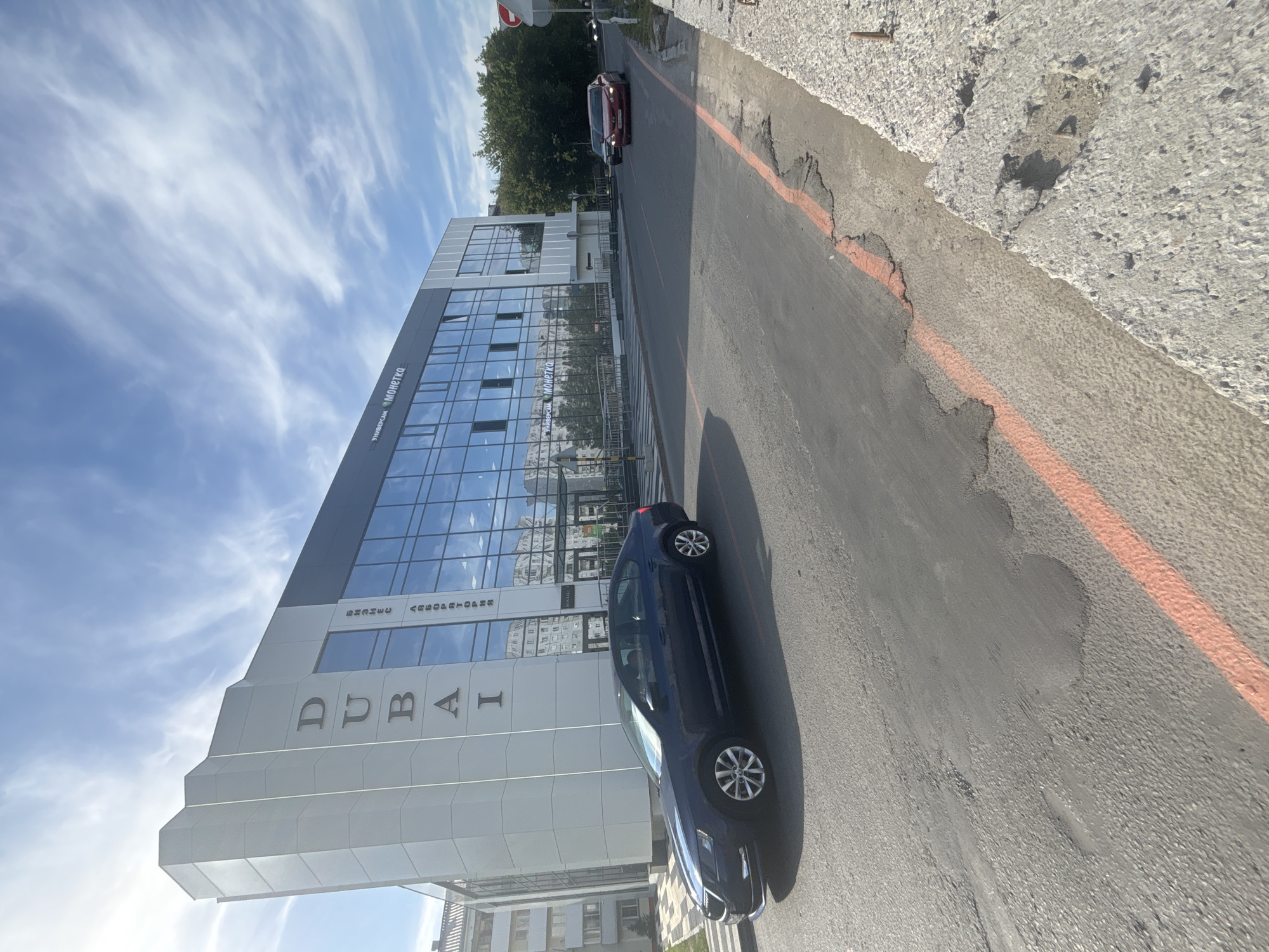 A dark blue sedan is parked on the right side of a road in front of a modern building with a glass facade. The building has the word "DUBAI" written vertically on its white facade. On the glass facade, Russian text is visible, including "БИЗНЕС АВТОРИЗАТОРИИ" and "УНИВЕРСАМ МОНЕТКА". The road has an orange marking on its right side, and further down the road, a red car is driving. The sky is blue with scattered clouds. This location is Novosibirsk, Russia.