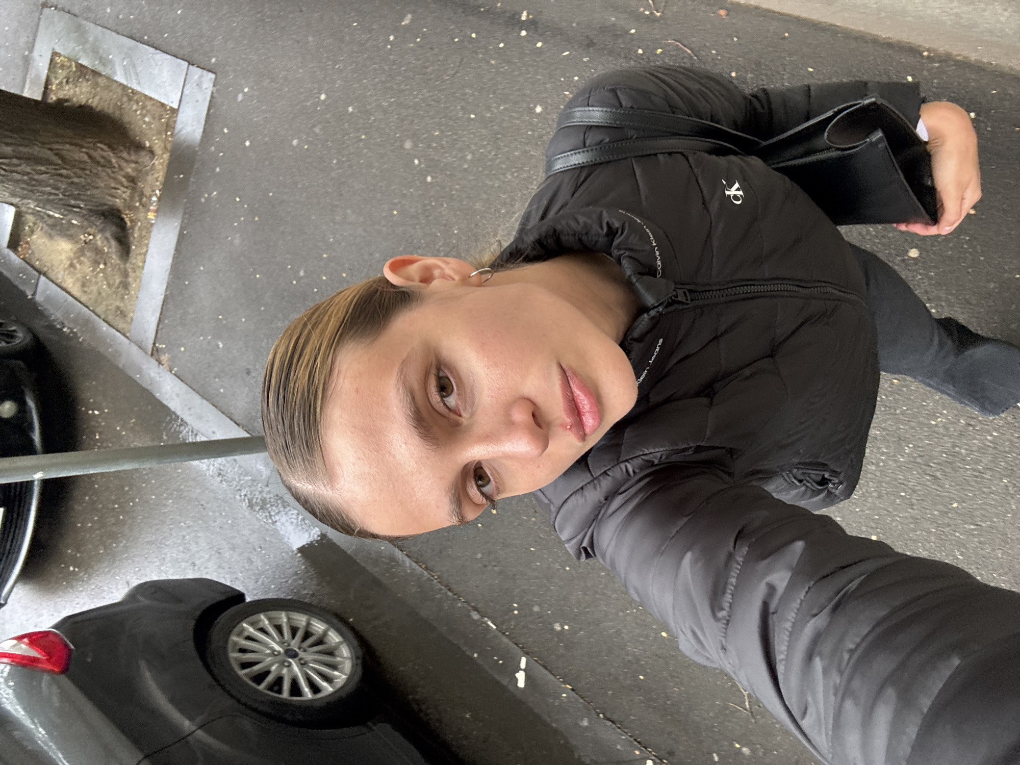 A young woman with light skin and light brown hair slicked back is captured in a selfie, angled slightly upwards towards her face. She wears a black puffer jacket with a visible "Calvin Klein Jeans" logo on the left chest and black pants. She is holding a black structured handbag with her right hand. A small silver hoop earring is visible on her left ear. Her expression is neutral, and her lips are slightly parted.
The background shows an outdoor urban setting. The ground is dark gray asphalt or concrete, appearing damp or recently wet, with scattered small light-colored specks. A black car is parked to her left, with its rear section, including a silver multi-spoke wheel rim (possibly a Ford, suggested by the wheel emblem visible in a crop), and part of a red taillight visible. A thin, dark metal pole stands vertically between the woman and the car. To her far left, the trunk of a tree emerges from a square, metal-edged planter box filled with dirt or mulch. The lighting is diffused, suggesting daytime, possibly overcast. The entire image is rotated 90 degrees clockwise from a standard upright orientation. The scene is consistent with a street or sidewalk in an urban area like Tbilisi, Georgia.