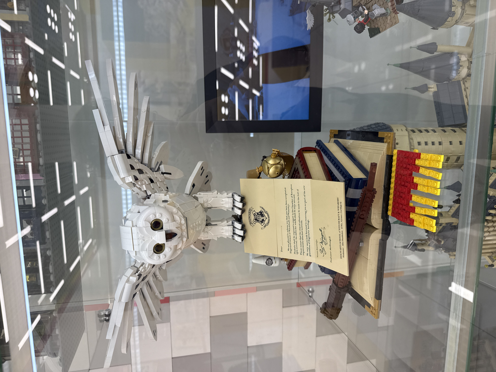 This is a close-up, eye-level shot of a Lego display inside a glass case, likely in a store in Khimki, Russia. The primary subject is a large, white Lego owl with outstretched wings, perched above a Lego replica of Hogwarts School. Below the owl is a Lego prop of an acceptance letter to Hogwarts, complete with the Hogwarts crest and a wax seal. The letter is placed on top of a stack of Lego books, and a Lego wand lies across the open book. To the right, a framed image is visible behind the Lego display, showing a blurry scene that appears to be another Lego creation or a photograph related to Harry Potter. The background of the display case is a reflective surface, showing the ceiling lights and a portion of the store. The overall scene depicts a detailed Lego model of iconic elements from the Harry Potter franchise.
