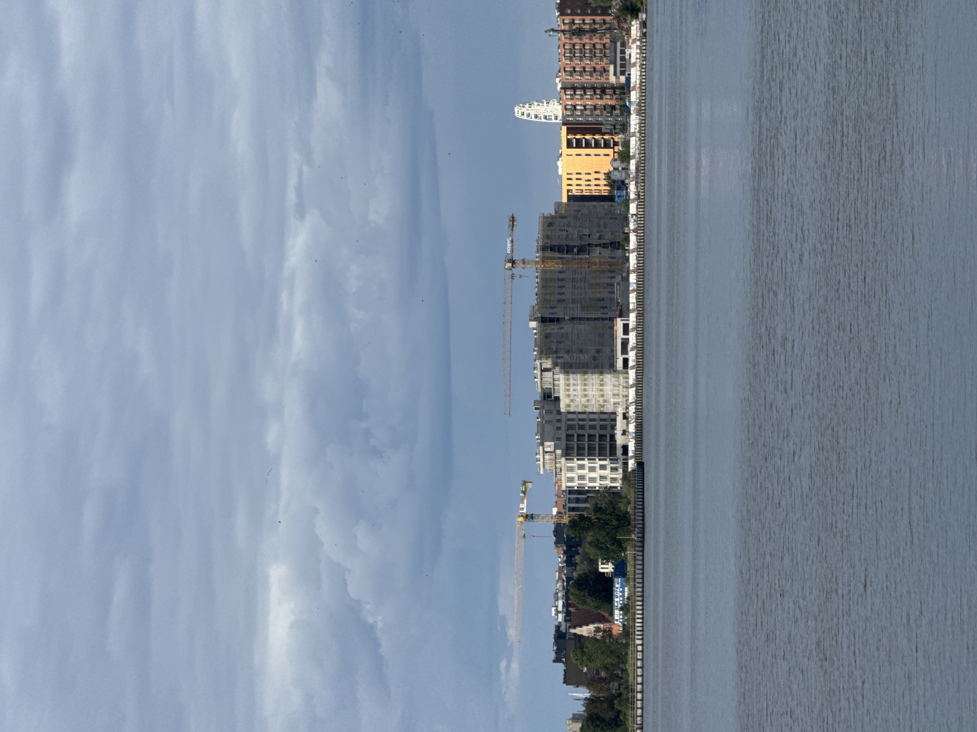 This is a full shot of the Gdańsk waterfront on a cloudy day. The foreground features a body of water with gentle ripples. In the mid-ground, a shoreline is visible with a line of moored boats or barges. Behind the shoreline, a row of buildings is under construction. Prominent among these are several large, multi-story buildings covered in scaffolding and protective netting, indicating ongoing construction. Two yellow construction cranes are visible, one extending horizontally and the other reaching upwards. Adjacent to the construction site are completed buildings, some with distinctive orange and brick facades. In the distance, a white Ferris wheel-like structure can be seen. The sky is filled with dramatic, grey clouds, with small birds scattered throughout the upper portion of the frame.