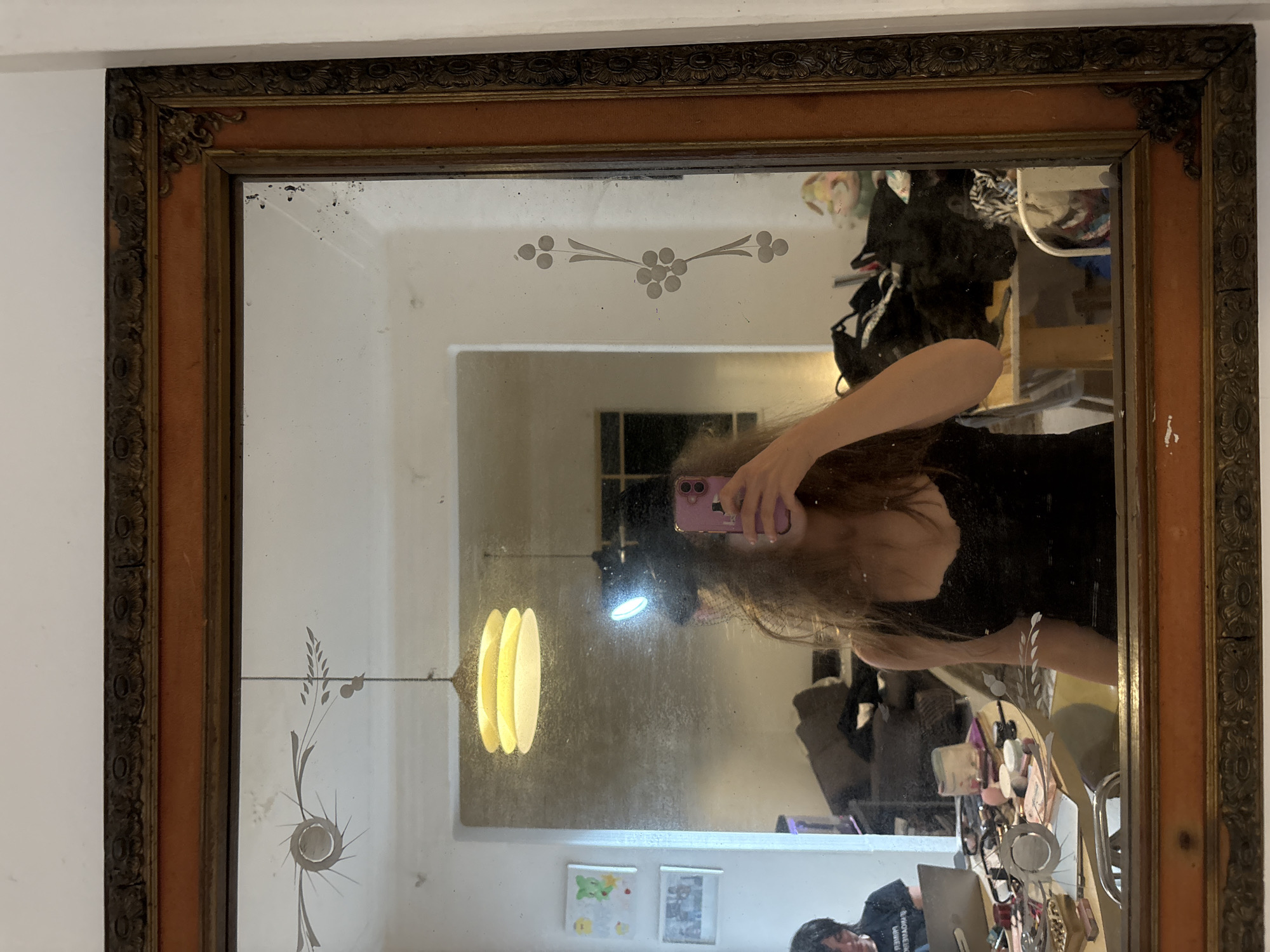 A framed mirror, likely located indoors in Mexico City, Mexico, reflects a person holding a pink cell phone, seemingly taking a selfie. The mirror has an ornate, aged frame with floral and leaf-like designs. The reflection reveals a cluttered room with clothes, furniture, and various personal items. The mirror itself has etched floral patterns at the top and bottom, and a pendant lamp is visible in the background. A collage of children's drawings hangs on the wall.