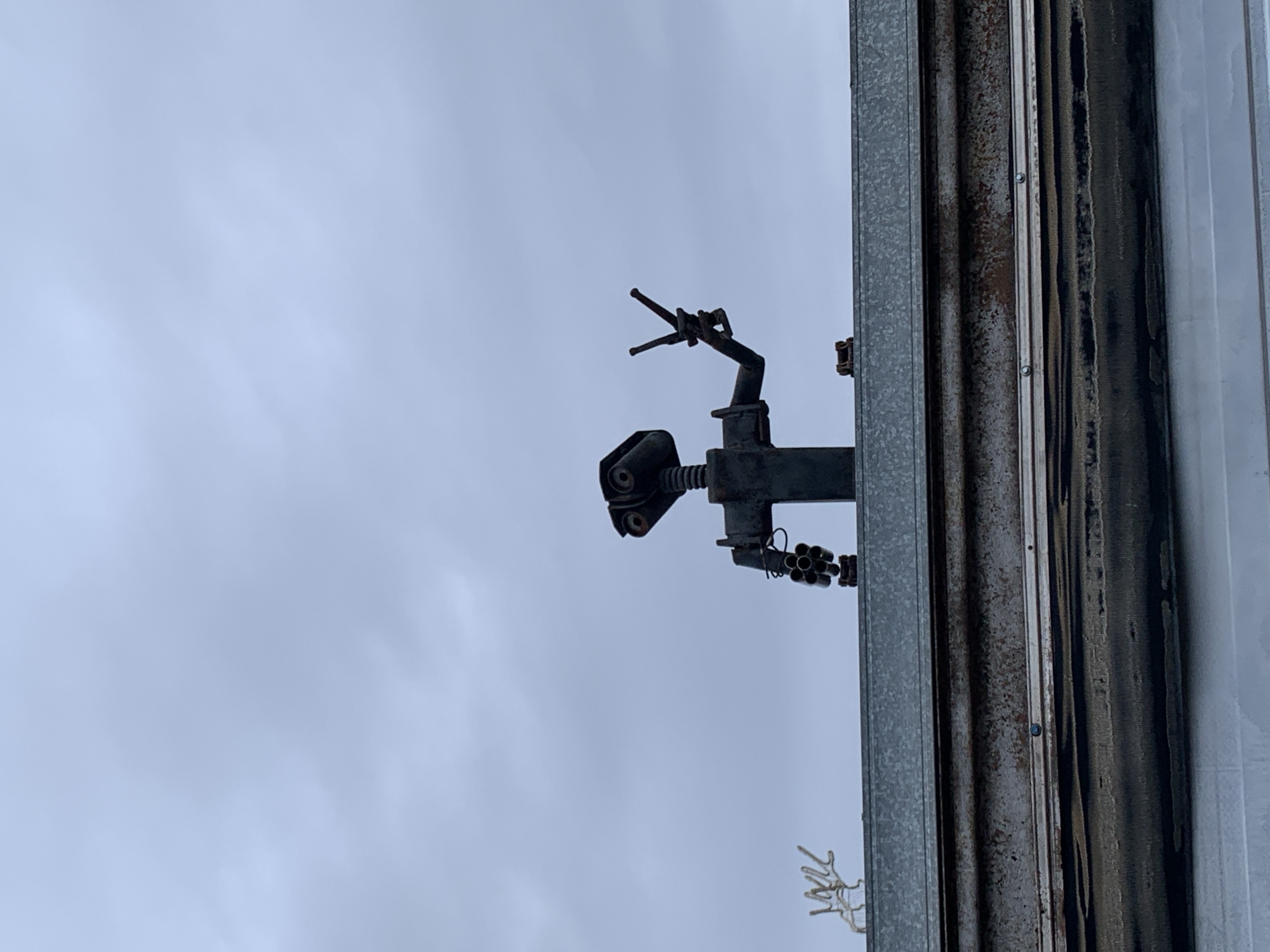 The image shows a metal sculpture of a deer head mounted on a metal building wall. The sculpture appears to be made of various metal components, including pipes, springs, and other mechanical parts. The building's wall is corrugated metal with some rust and discoloration. The sky is overcast. There is no evidence to confirm the location in Фоки, Russia.