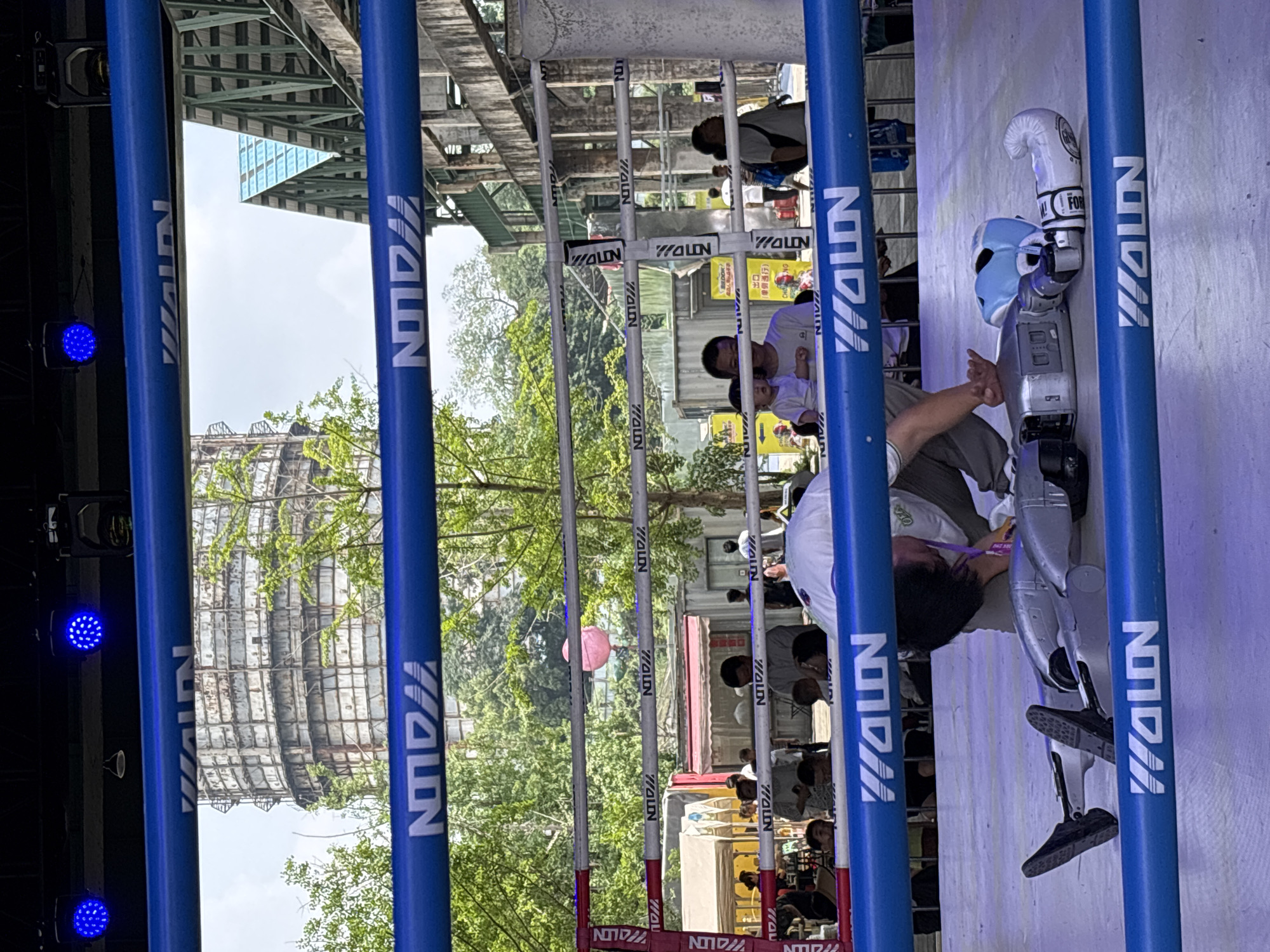In the city of Dadukou, China, a boxing ring is set up outdoors, with blue ropes and posts. Inside the ring, a person wearing a white and purple outfit, appears to be in a prone position, possibly injured or exhausted, near a silver robot. A blue boxing helmet and a white boxing glove are also on the canvas. The background shows trees, a large industrial structure, and a crowd of onlookers outside the ring. The scene suggests an event involving robots and possibly combat sports.