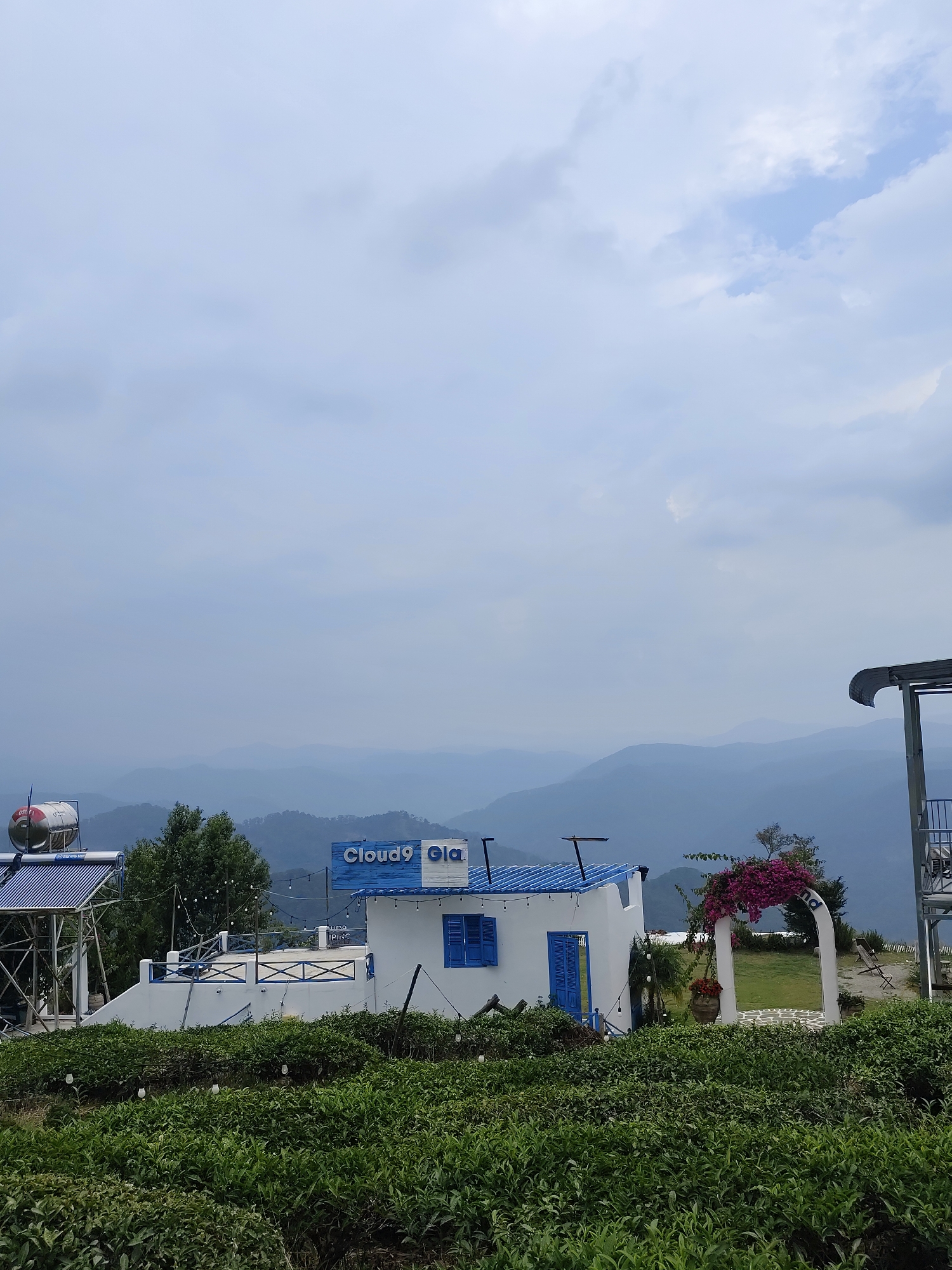 The image depicts a landscape in Phát Chi, Vietnam, featuring a white building with blue accents, possibly a café or guesthouse named "Cloud9 Gla," set against a backdrop of hazy, distant mountains. In the foreground, a dense expanse of green tea bushes occupies the lower portion of the frame. To the left of the main building is a structure with a solar water heater. An arched structure decorated with pink flowers sits to the right of the building. The sky is overcast with varying shades of gray.