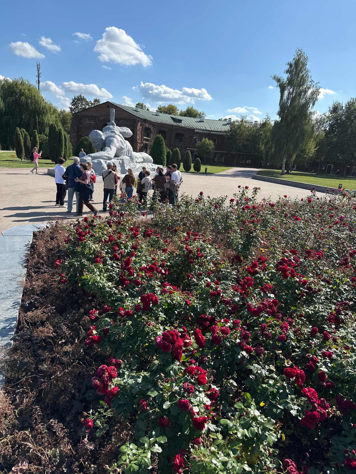 A group of people are gathered in front of a large, light-colored sculpture of a fallen soldier in the city of Brest, Belarus. The sculpture is situated in a paved area with a large flowerbed of red roses in the foreground. In the background, there is a red brick building with a green roof, and several trees. The sky is blue with white clouds. The people appear to be tourists, and some are taking photographs. One person in a pink shirt is walking away from the group and taking a picture with their phone.