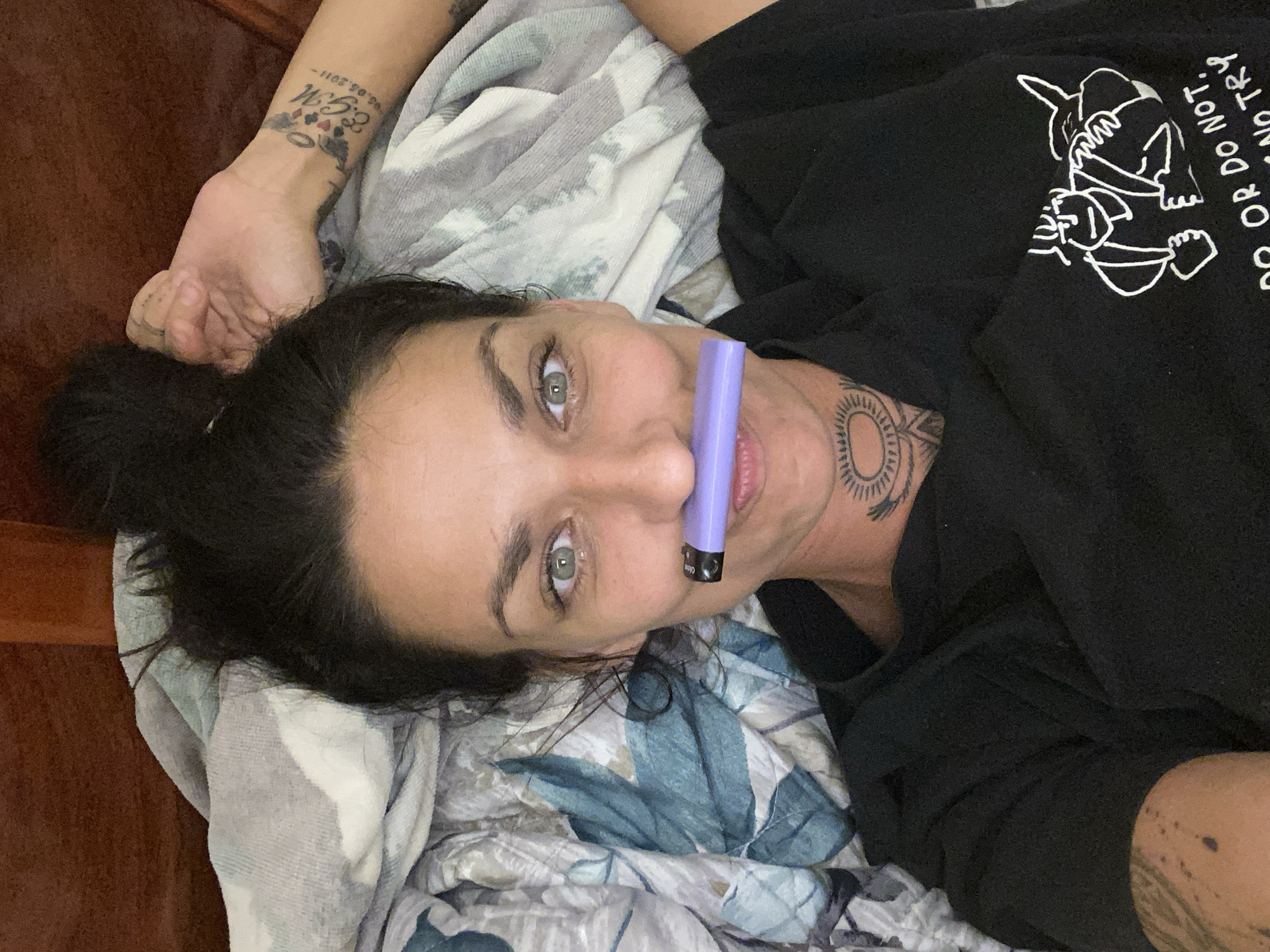 A woman lies in bed, holding a purple vape pen to her mouth. She has dark hair pulled up in a bun and green eyes. She is wearing a black t-shirt with a graphic design on it and has tattoos on her neck, arm, and wrist. The background is a floral comforter and a wooden headboard. The image is a close-up selfie.