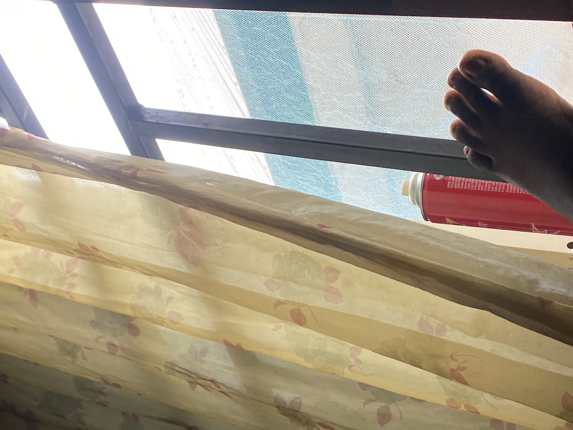 In Jauro Yinu, Nigeria, a close-up shot shows a person's bare foot, with toes curled, positioned near a window. The foot is silhouetted against bright light filtering through a sheer, yellow curtain with a floral pattern. To the left of the foot, a red spray can is visible, partially obscured by the curtain. The window is framed by dark vertical bars and covered with a fine mesh screen, through which a bright sky and distant buildings are faintly discernible.