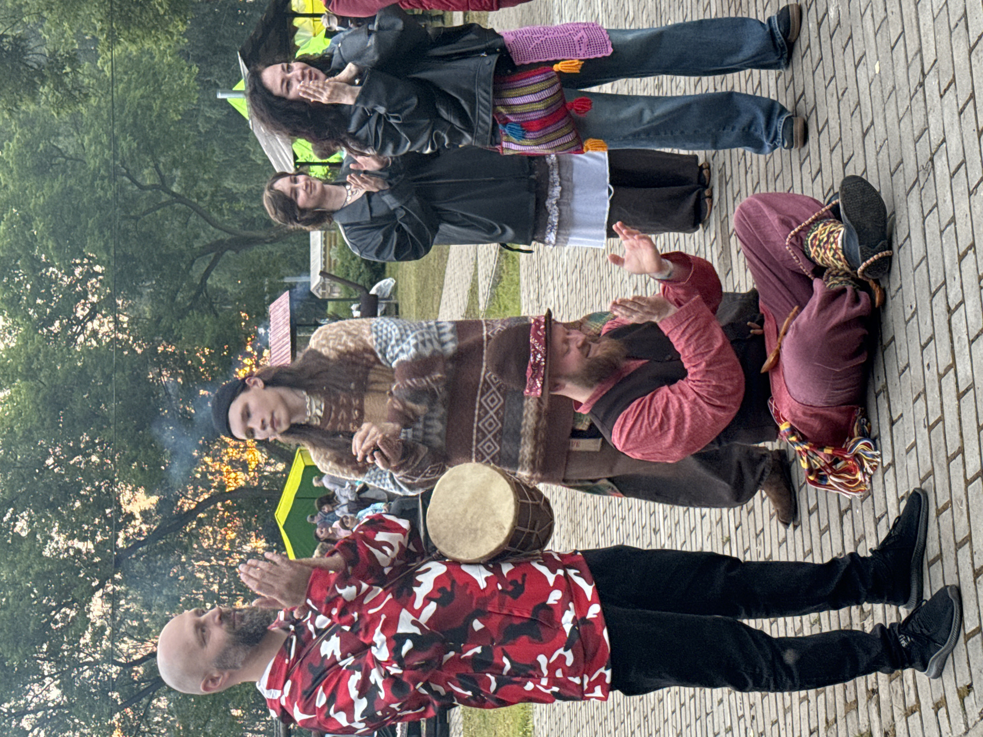 In Старые Быги, Russia, a group of people are gathered outdoors on a cobblestone path, possibly for a festival or event. The scene is bathed in the warm glow of sunset, suggesting it is late afternoon or early evening. The trees in the background are full of green leaves, and the sky appears to be clear.
The individuals in the foreground are casually dressed, many in bohemian-style clothing. One man in a red and black camouflage shirt is clapping his hands, looking upwards with a serene expression. Beside him, another man with a beard is looking down at a small drum placed on his lap. A woman wearing a patterned blanket-like garment is also clapping her hands. Further back, two women are visible, one in a black leather jacket and the other in a dark jacket, both with their hands clasped together. Another person in maroon pants and a pink top is seated.
The overall atmosphere seems relaxed and celebratory, with a sense of communal engagement suggested by the shared activity of clapping and the presence of musical instruments like the drum. The setting, with its natural elements and the paved walkway, hints at an outdoor gathering space. No discernible text is visible in the image.