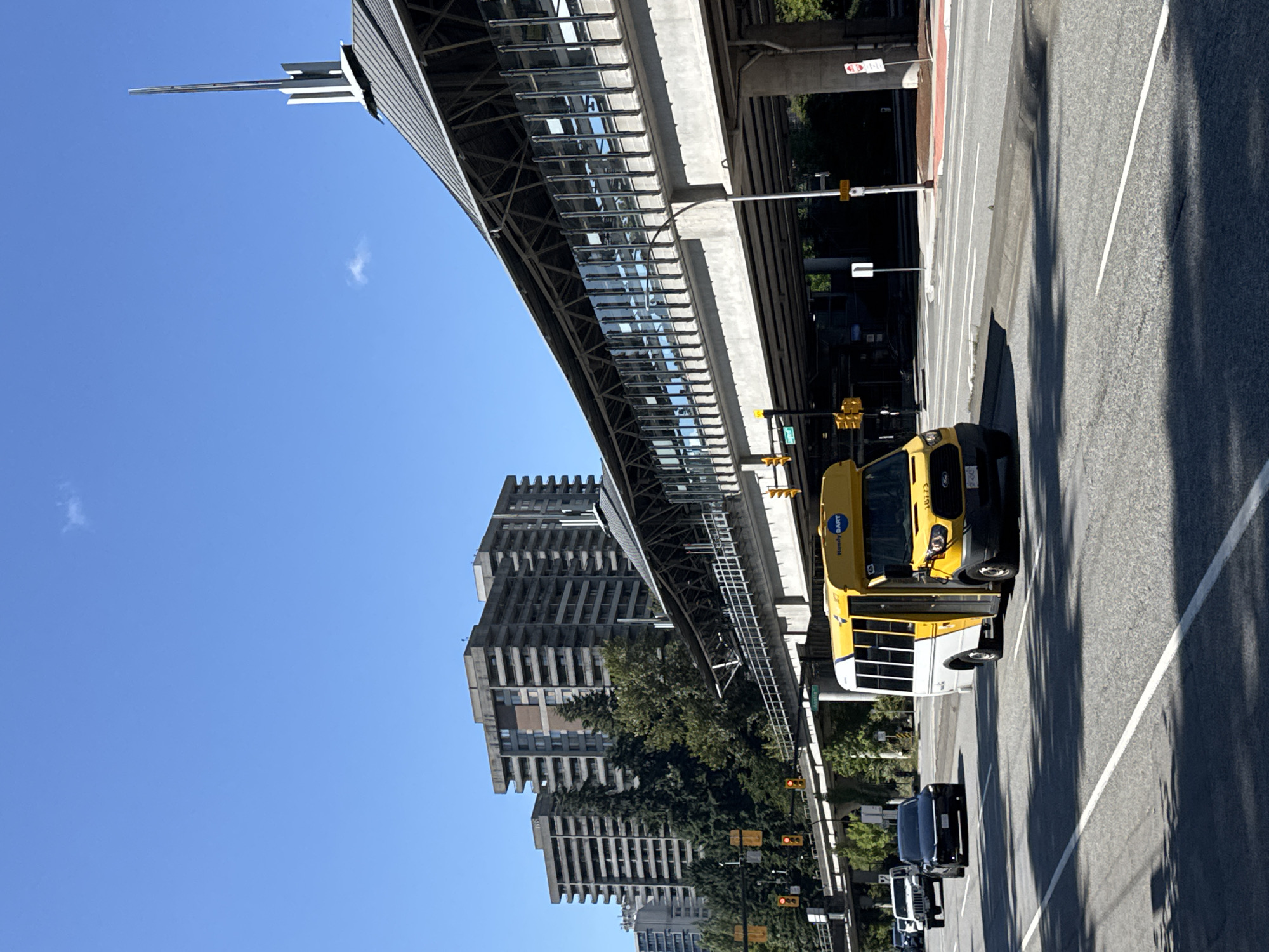 An outdoor, eye-level, medium shot captures a sunny daytime scene on a street in Burnaby, Canada. In the foreground, a yellow and white minibus with "Handy DART" branding is driving on the road, followed by a black sedan and a white Jeep. The road is paved with asphalt and has white lane markings, with shadows of trees cast across it.
In the midground, a modern elevated transit station with a grey roof and glass walls runs diagonally across the frame. Beneath the station, an elevated guideway, likely for an automated train system, is visible. Traffic signals with yellow and red lights are mounted on poles near the intersection.
In the background, several tall, brutalist-style apartment buildings with many balconies dominate the skyline. Lush green trees are interspersed between the buildings and the transit structure. The sky is clear and bright blue with a few small, wispy clouds. The overall atmosphere is bright and urban, suggesting a busy transit corridor within the city.
