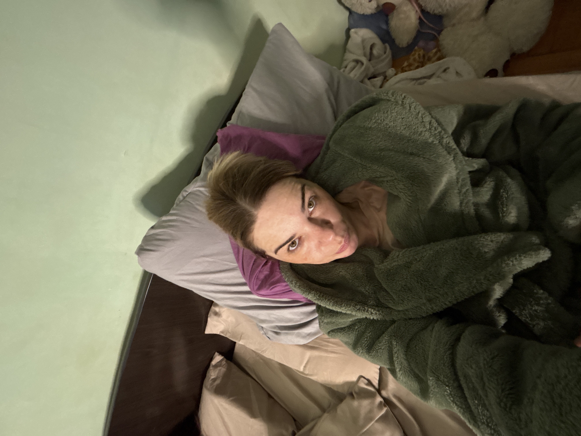 A medium, high-angle shot depicts a person lying in bed. The individual has fair skin, light brown hair, and is gazing upwards towards the camera. They are wrapped in a green, fluffy robe. The bed is layered with a grey pillow, a purple pillow, and beige bedding. In the background, partially visible, are plush toys. The scene appears to be indoors, likely a bedroom, with a light green wall visible on the left. The location is unknown.