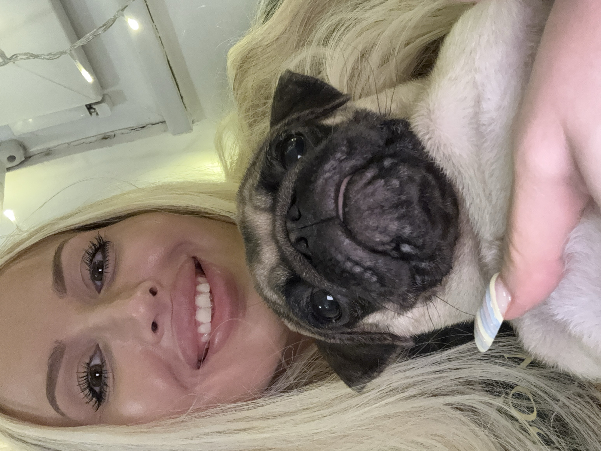 A close-up, selfie-style image shows a blonde woman and a pug puppy. The woman is smiling widely, with her eyes looking directly at the camera. She has long blonde hair and is wearing makeup. The pug puppy is positioned in front of the woman, with its face filling a significant portion of the frame. The puppy has dark markings around its eyes and muzzle, and its tongue is slightly visible. The background is blurred but appears to be indoors, with some string lights visible, suggesting a festive or cozy environment. The lighting is bright and appears to be artificial.