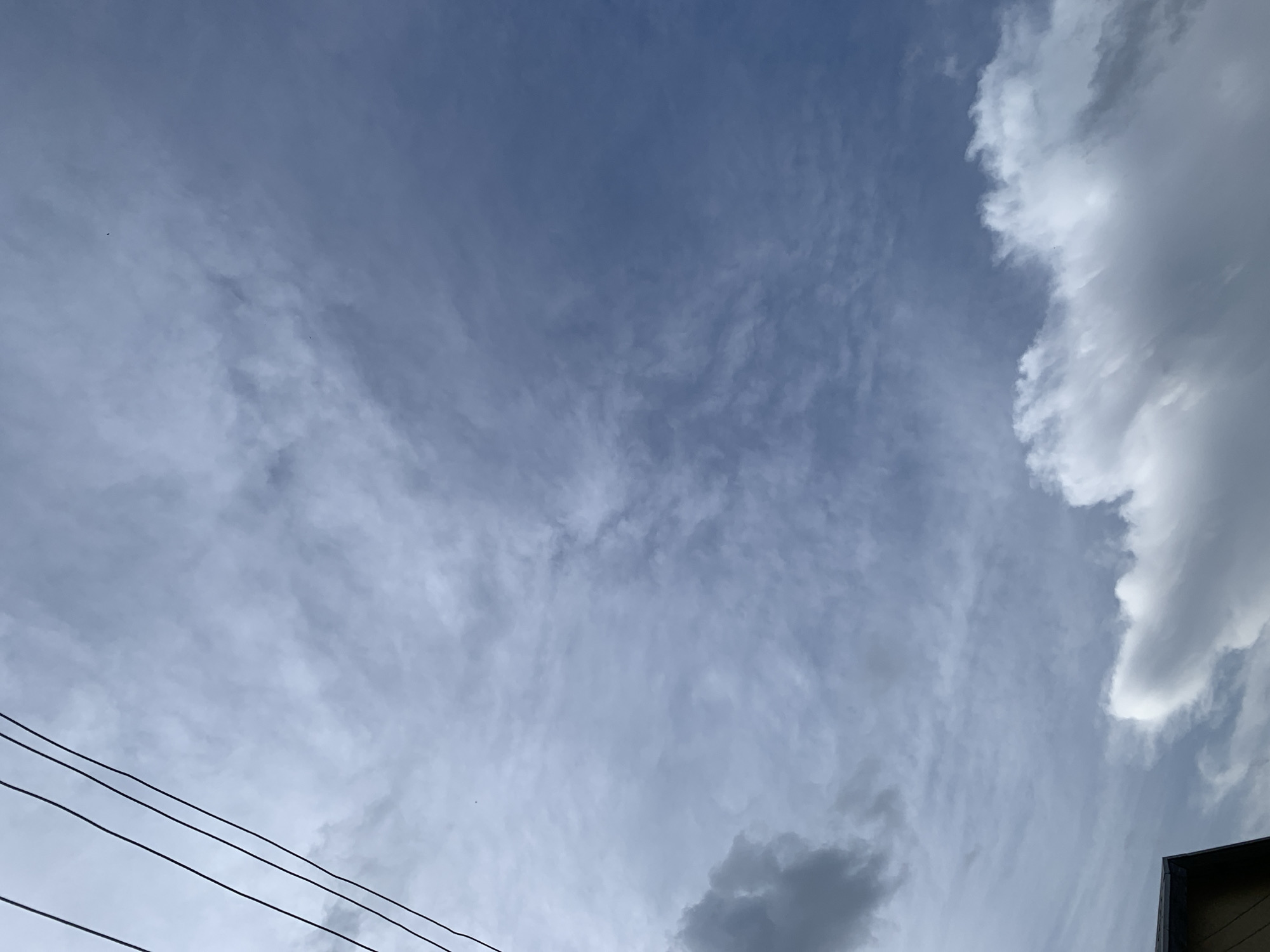 The image depicts an outdoor scene dominated by a cloudy sky. The sky is a muted blue, with wispy, cirrus-like clouds stretching across much of the frame. On the right side of the image, larger, fluffier cumulus clouds are visible, some appearing dense and white, while others are darker and more shadowed, suggesting varying cloud formations and possibly impending weather changes.
In the lower left portion of the image, three parallel, dark lines traverse the frame horizontally. These appear to be power lines or telephone wires, indicating a man-made presence in the otherwise natural sky. In the far lower right corner, a sliver of what appears to be the roofline of a building is visible, painted in earthy tones, further grounding the image in a terrestrial setting.
There are no people visible in the image. The activity taking place is simply the natural movement and formation of clouds in the sky. The time of day is not explicitly clear, but the lighting suggests it is daytime, likely mid-morning or afternoon, given the presence of sunlight illuminating the clouds. The weather appears to be dynamic, with a mix of clear sky, wispy high clouds, and more substantial cumulus clouds, possibly indicating a day with changing weather conditions. The presence of power lines and a building suggests an inhabited or developed area, possibly a residential or urban environment. No text is visible in the image.