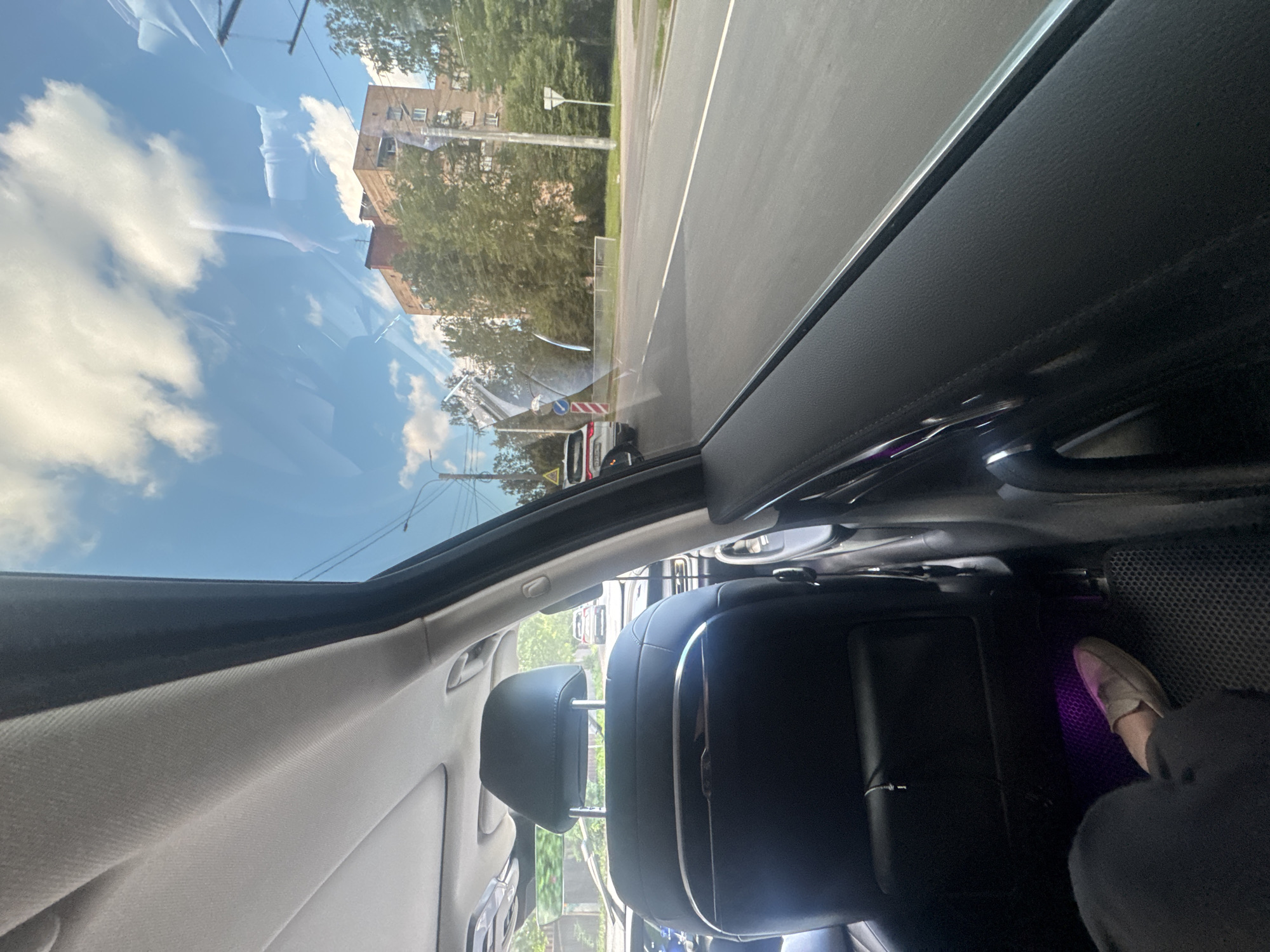 The image shows the interior of a car, looking out the side window. The back seats of the car are visible, as well as a person's leg and foot on the floor. Outside the window, a street scene is visible with buildings, trees, and other cars. The sky is blue with some clouds. The image appears to have been taken during the daytime.