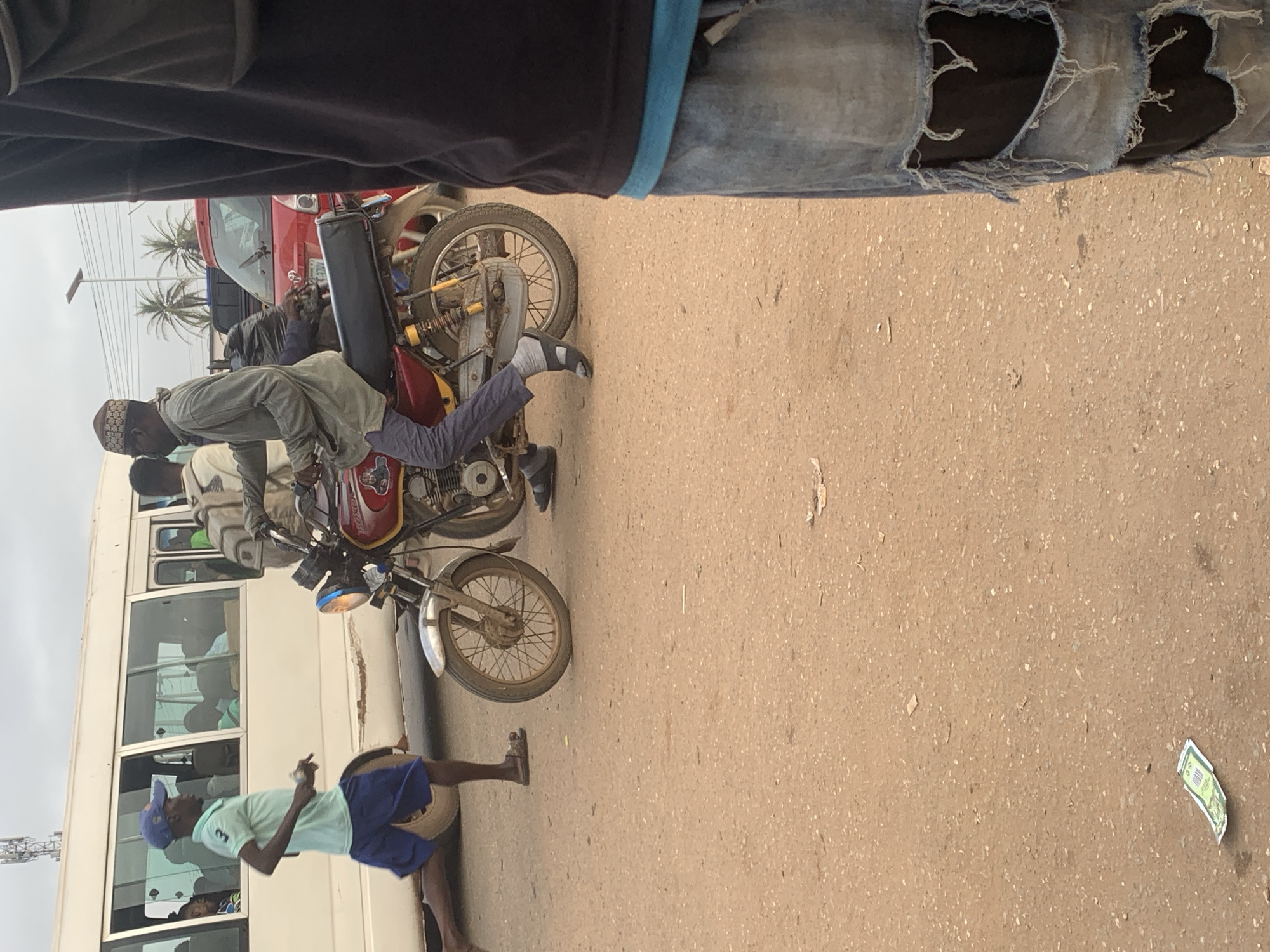 In Shagamu, Nigeria, a man wearing a patterned cap, a light grey long-sleeved shirt, and grey pants rides a red motorcycle. He is wearing dark sandals over light grey socks. The motorcycle has a yellow stripe along its side and a black seat. The man appears to be looking forward. To the left of the motorcycle, a young man in a light blue short-sleeved shirt, blue shorts, and a blue cap runs alongside a white bus. He is wearing dark sandals. The bus has several passengers visible through its windows. In the foreground, on the right side of the image, a person wearing ripped blue jeans and a black shirt stands partially visible. The ground is a dusty, reddish-brown color, characteristic of roads in the area. A green banknote is visible on the ground to the left of the motorcycle. Palm trees and power lines are visible in the background, with a red car behind the motorcycle. The sky is overcast.