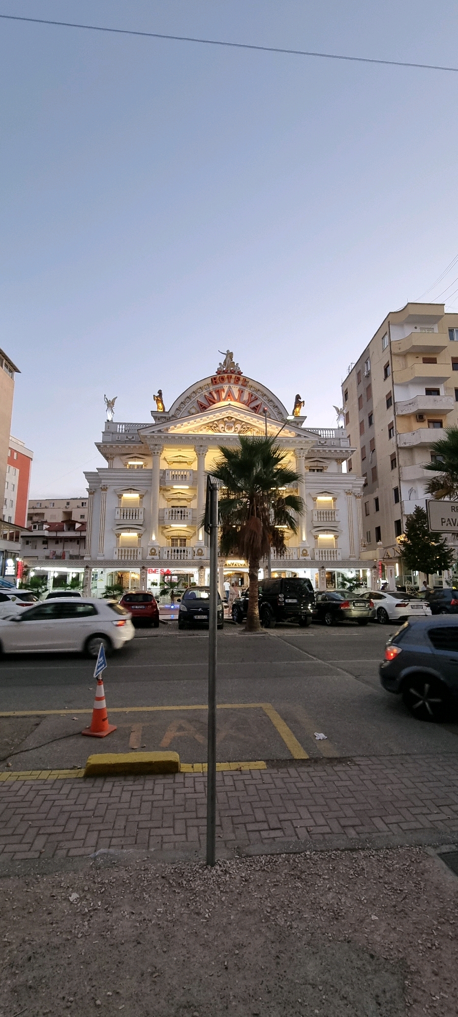 The image shows a street scene in Bashkia Durrës, Albania, featuring the Hotel Antalya. The hotel is a prominent, white, multi-story building with ornate architectural details, including columns, balconies, and statues. A large palm tree stands in front of the hotel, and other buildings, including a taller apartment building, are visible to the right and left. Several cars are parked and in motion on the street in front of the hotel and in a parking area. A traffic cone and a pole with a sign are in the foreground. The sky is a clear, light blue, indicating either early morning or late afternoon. The street is paved, and there is a yellow line marking a parking or traffic lane.