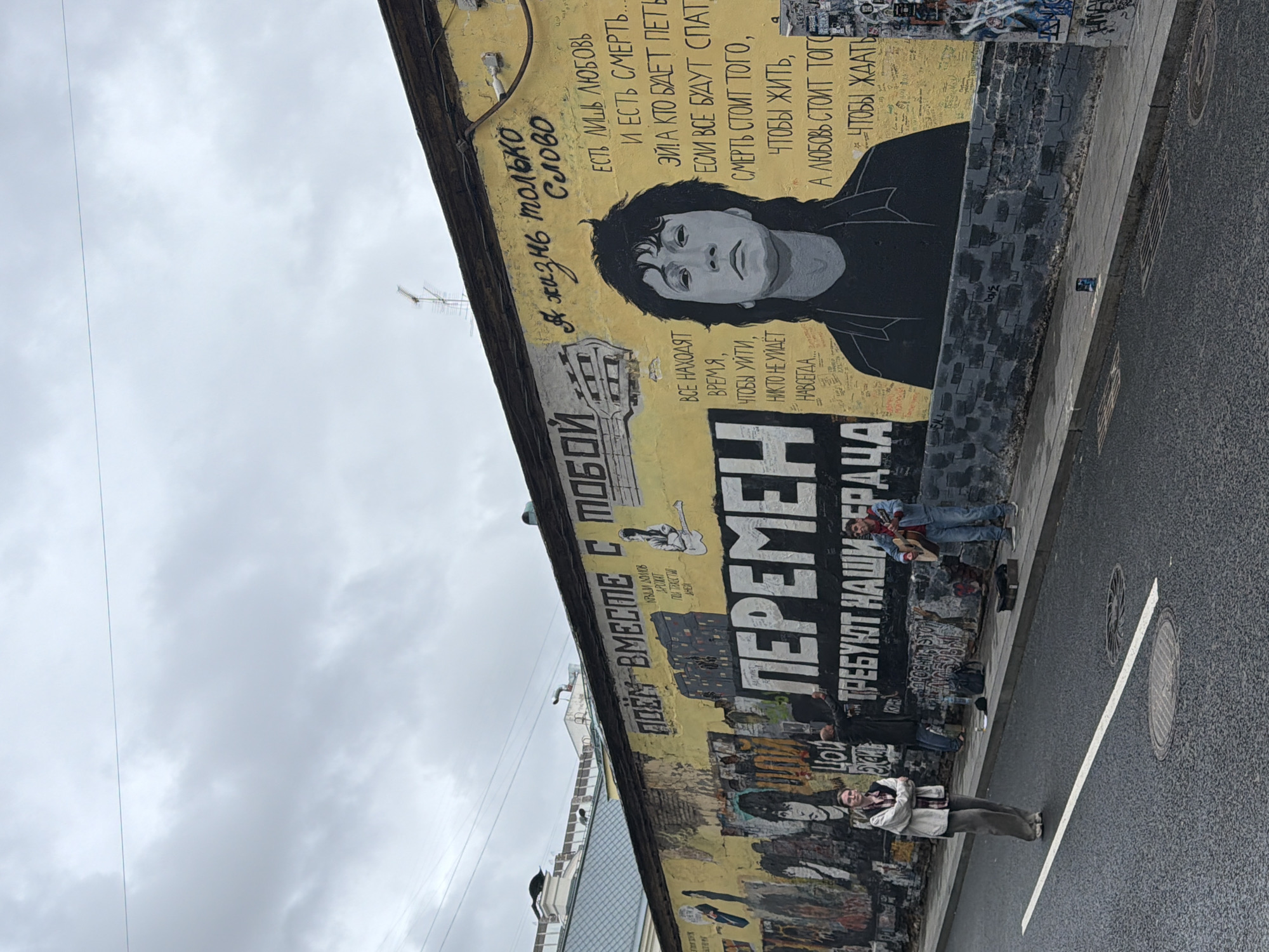 This is a photograph taken from a slightly elevated, angled perspective, looking down at a street scene dominated by a large, yellow mural on a building wall. The mural features a portrait of a person with dark, wavy hair, rendered in grayscale, looking directly at the viewer. To the left of the portrait, Russian text is written vertically in black. Below the portrait, more Russian text appears in large, bold, black letters, forming the word "ПЕРЕМЕН" (PEREMEN), which translates to "CHANGE". To the right of this word, smaller black Russian text is visible, reading "ТРЕБУЮТ НАШИ" (TREBUYUT NASHI), meaning "DEMAND OUR".
Several people are present in the scene. In the upper right portion of the mural, two figures are depicted on the wall itself, one appearing to play a guitar. On the sidewalk below the mural, three people are visible. One person is lying down on the sidewalk, while another is sitting with their legs extended. A third person is standing further down the street. In the foreground, a person is standing on the wet asphalt, looking towards the building. The weather appears overcast, suggesting a cloudy day. The street is paved with asphalt and includes a manhole cover with a circular pattern. A white line is painted diagonally across the street. The overall mood seems somber, possibly due to the overcast weather and the themes implied by the mural's text and imagery.