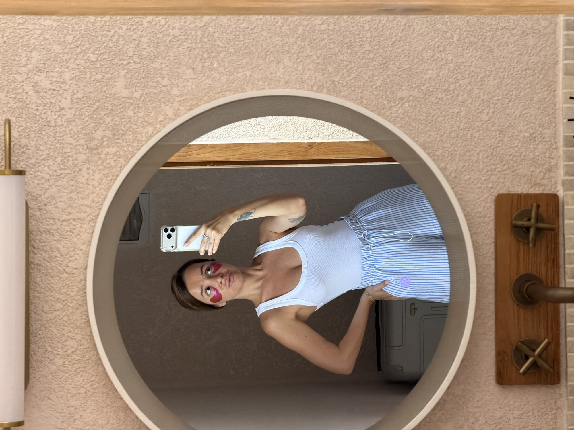 A young woman with brown hair and heart-shaped eye patches is taking a selfie in a circular mirror in what appears to be a hotel bathroom in Mengwi, Indonesia. She is wearing a white tank top and blue and white striped shorts. Her left arm is on her hip, and she is holding a smartphone in her right hand. The mirror reflects a portion of the room behind her, including a suitcase and a doorway. The bathroom walls are a textured pinkish-beige color. A wooden panel with brass fixtures is visible below the mirror, suggesting a sink area.