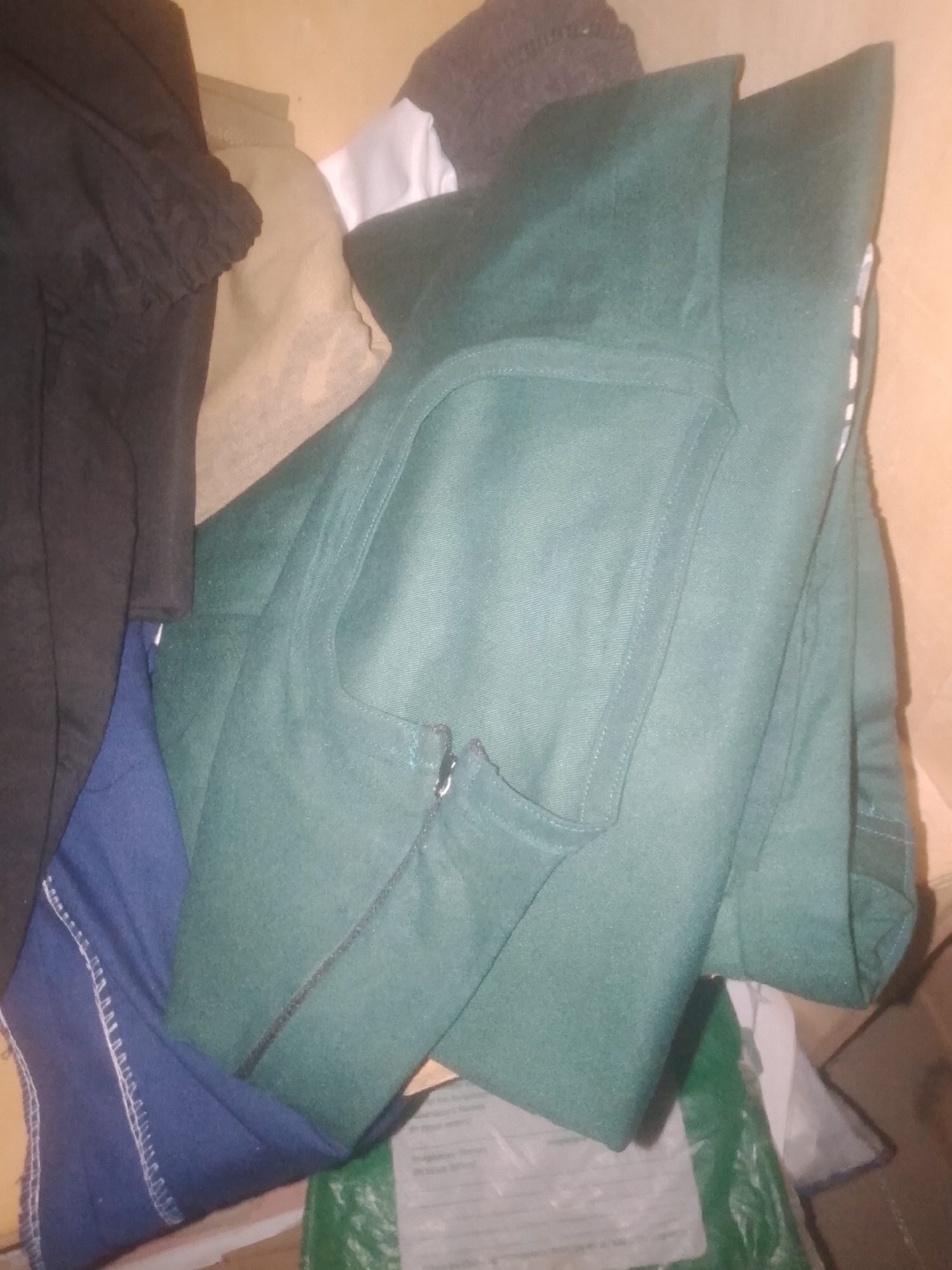 This is a close-up, high-angle shot of a pile of clothing items. The dominant item is a dark green garment, possibly a jacket or a coat, folded in half. It features a prominent stitched pocket on the right side and a zipper closure running diagonally. To the left of the green garment, a portion of a black garment, likely a sleeve with ruffled detail, is visible. Beneath these, a dark blue fabric with visible white stitching along its edge is seen, possibly part of a pair of pants or another garment. In the background, hints of other fabrics are present, including a tan-colored material and a small section of white fabric, along with what appears to be the edge of a dark grey knitted item. The lighting is warm and uneven, suggesting an indoor setting with artificial light. There is no identifiable background that would indicate a specific location within Jauro Yinu, Nigeria, or any other city. The image focuses solely on the textures and colors of the folded clothing.