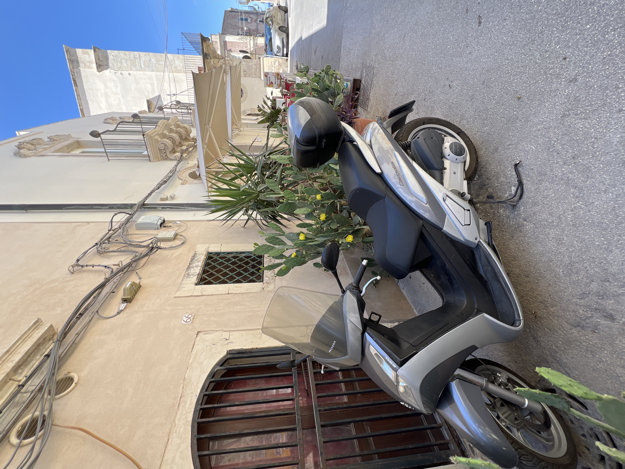 A silver Peugeot scooter is parked on a paved street in front of a beige building in Syracuse, Italy. The building has brown metal bars on a doorway on the left, a small barred window above it, and an ornate balcony with intricate stonework on the upper floor. To the right of the scooter, there is a cluster of prickly pear cacti with yellow flowers and a light-colored awning extending from the building. Further down the street, a white car is visible, and the sky is clear and blue. The scooter has a large black storage box on the rear seat and a tinted windshield. The scene appears to be in a narrow street with a slightly inclined road.