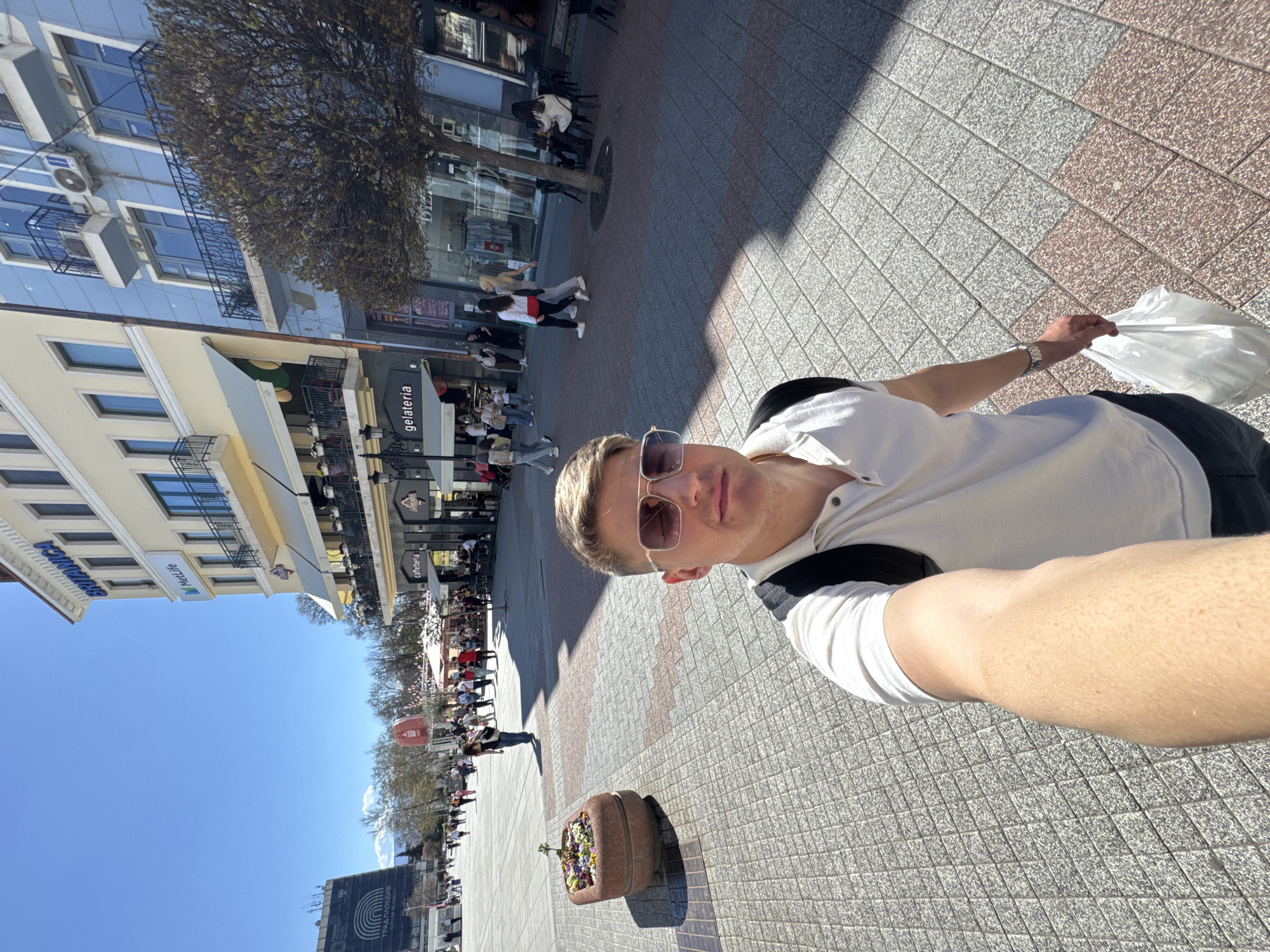 A man takes a selfie on a sunny day in Plovdiv, Bulgaria. He is wearing sunglasses and holding a white plastic bag. The background shows a pedestrian street with various cafes, shops, and people walking and sitting. Buildings with apartments are visible on the left side of the frame. The ground is paved with gray and red tiles. A flower pot sits in the foreground. A sign in Cyrillic and Latin alphabets is visible in the far background.