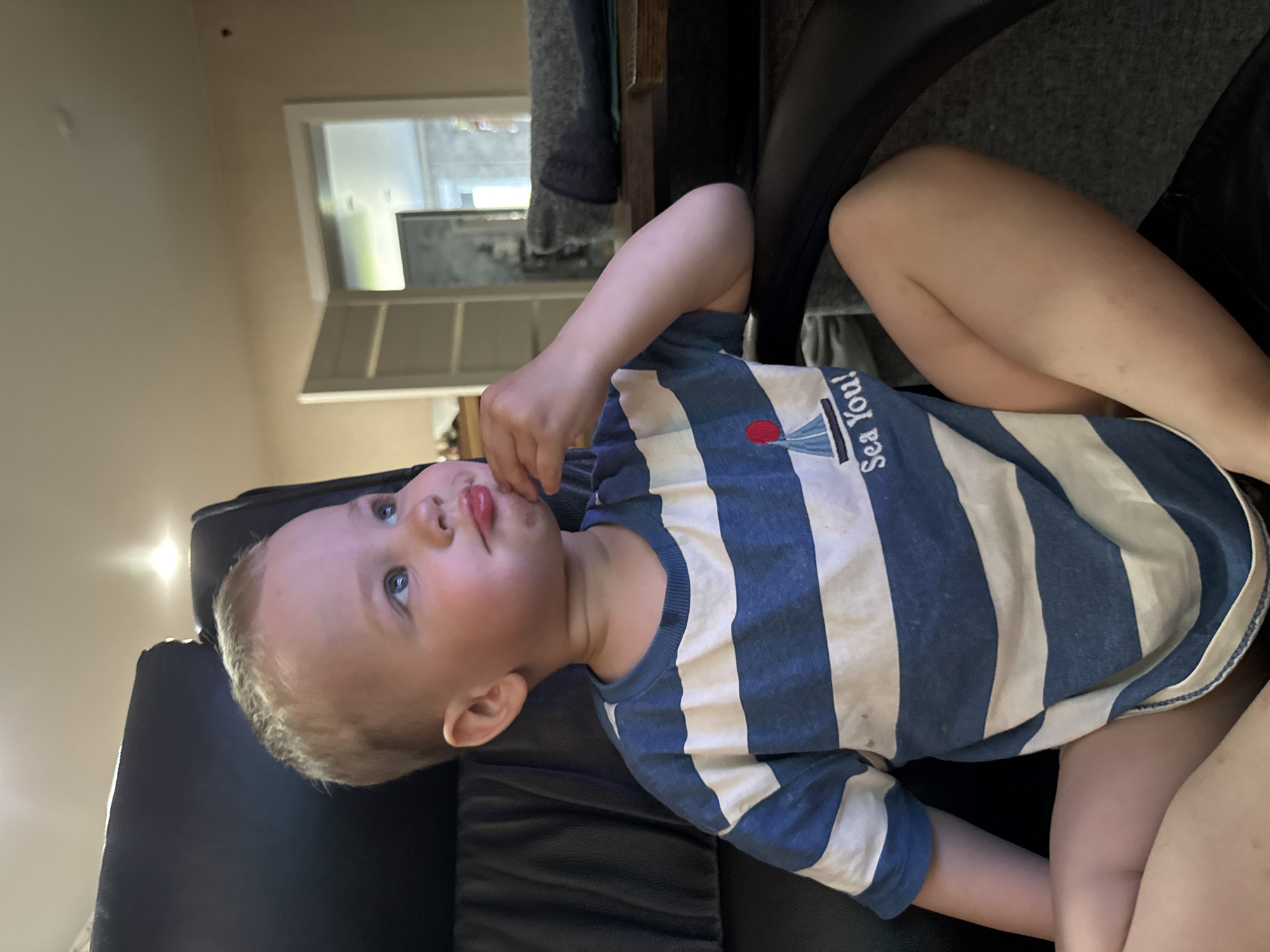 A young child is sitting in a black chair, possibly indoors. The child is wearing a blue and white striped shirt with a graphic of a sailboat and the words "Sea You!" on it. The child's head is turned upwards, and they appear to be touching their mouth with their fingers. The background shows a glimpse of a doorway and a window. The location is Unknown, Unknown.