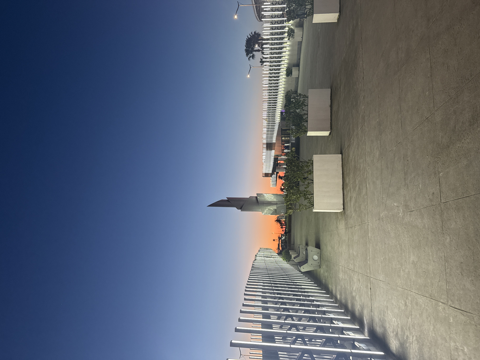 The photograph depicts a view in Batumi, Georgia, at dusk. The sky transitions from deep blue to orange near the horizon. A modern architectural structure with a pointed apex stands prominently against the colorful sky. A long row of illuminated, linear light fixtures extends from the bottom of the frame towards the center, adjacent to a paved area. White planters containing greenery are spaced along the paved surface. Palm trees are visible in the background. The scene lacks visible people.