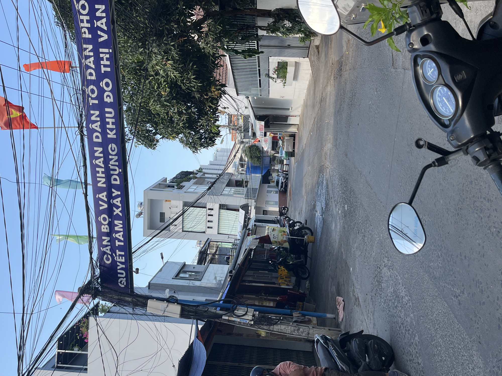 A street in Bắc Nha Trang Ward, Vietnam, is seen from the perspective of a parked scooter. The street is lined with buildings of varying heights, some under construction, and motorbikes parked along the side. A blue sign with white Vietnamese text hangs over the street, supported by a web of electrical wires. Flags of various colors hang from the sign. The sky is clear and blue. The scene has a bright, sunny quality.