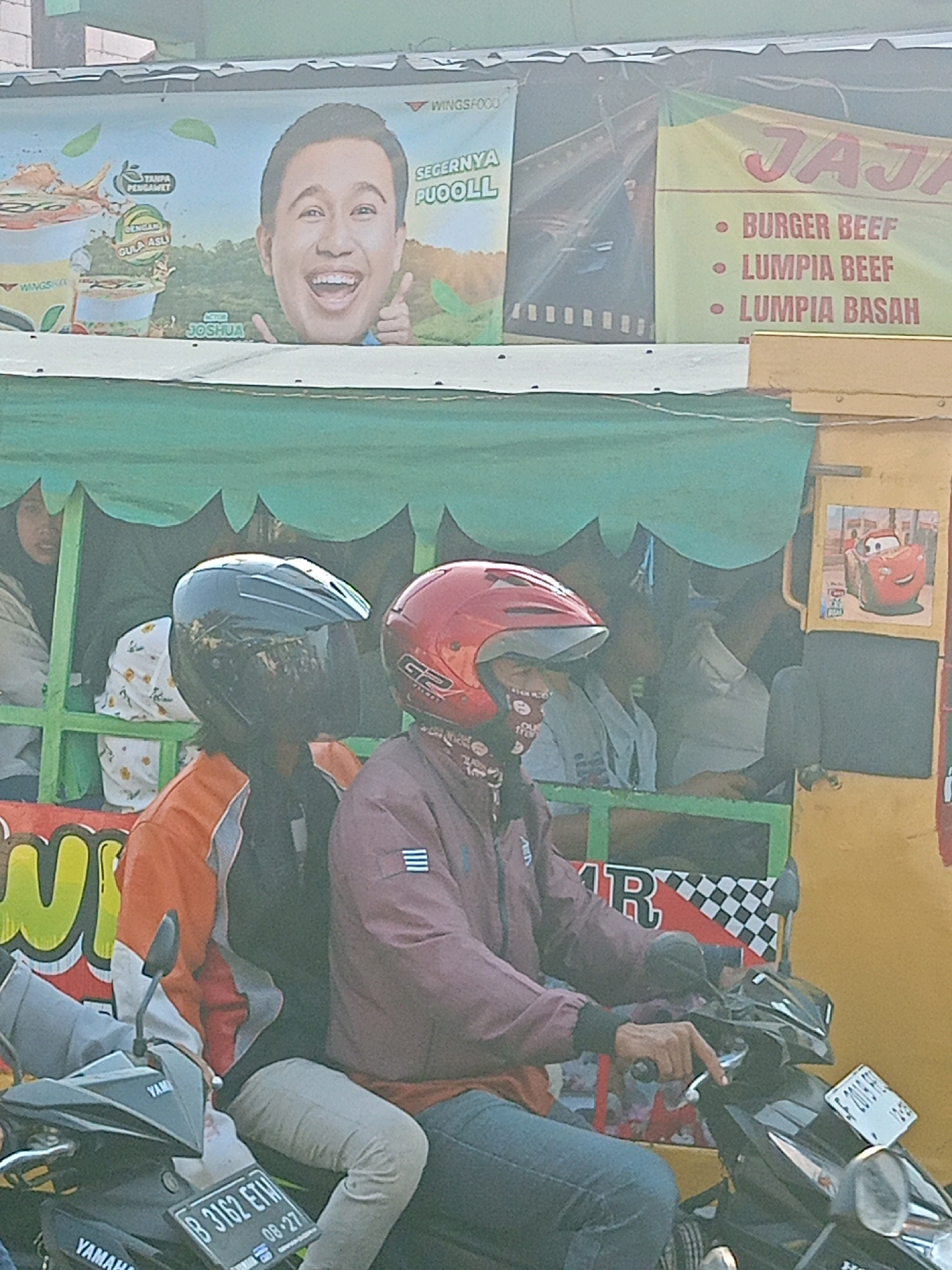 The image depicts a street scene in Satriamekar, Indonesia, where two individuals, possibly a couple, are seated on a motorcycle. The person in the front, wearing a maroon jacket and jeans, has their right hand on the motorcycle's handlebar. The person behind them, in an orange and black jacket, is partially obscured. Both are wearing helmets, with the front rider sporting a red helmet and the rear rider a dark blue helmet.
The motorcycle is parked in front of a stall or food cart, indicated by the colorful banners and awnings. One banner features a large headshot of a man and text that includes "SEGERNYA PUOLL" and "ACTOR JOSHUA". Another banner to the right displays food items like "BURGER BEEF", "LUMPPIA BEEF", and "LUMPPIA BASAH". A cartoon image of Lightning McQueen from the movie Cars is also visible on the side of the cart. The background suggests an outdoor, urban environment. The image is taken from a slightly elevated angle.