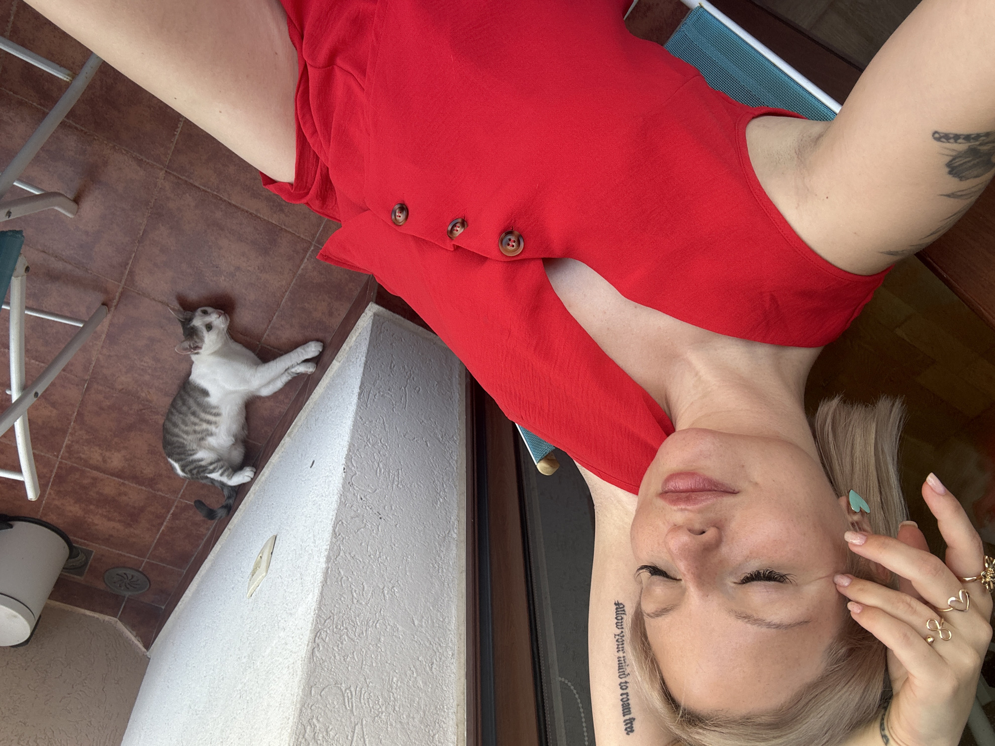 A high-angle, full-body shot captures a woman lying on a tiled patio outdoors, possibly in Budva, Montenegro. She is wearing a vibrant red romper. Her eyes are closed, and her head is tilted back, resting on a wooden surface. Her right hand is near her face, adorned with rings, and her left hand is on her chest. A cat with grey and white markings lies relaxed on the tiled floor to her left, looking upwards. In the background, parts of a chair and a trash can are visible. The lighting suggests it is daytime, with natural light illuminating the scene. A tattoo is visible on her left forearm with the text "Allow your mind to roam free." Another tattoo is on her upper arm. The woman appears to be in a state of relaxation.