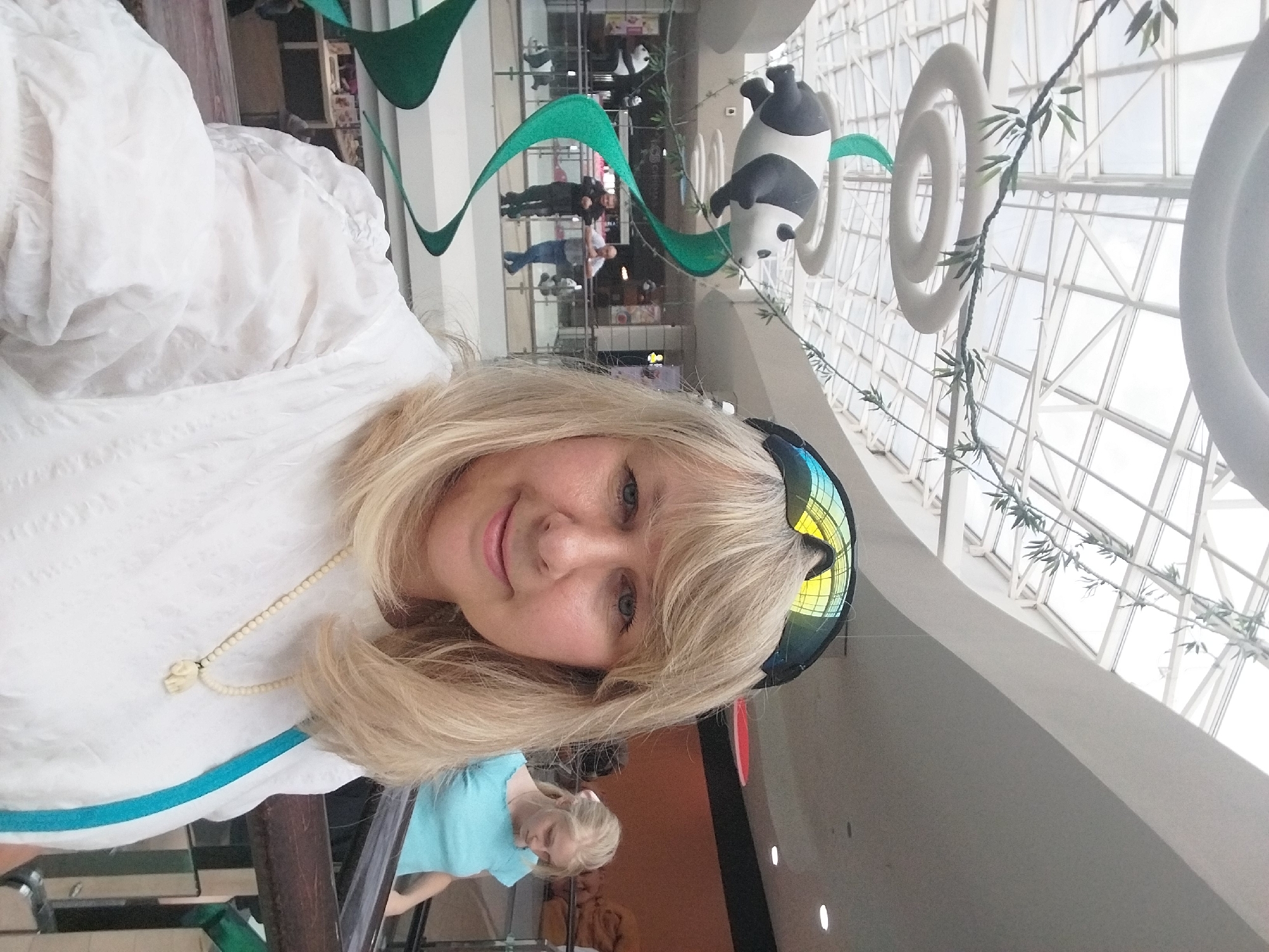 The image depicts a woman taking a selfie in what appears to be a shopping mall. She has blonde hair, blue eyes, and is wearing a white textured top and a necklace with an elephant pendant. She is also wearing sunglasses on top of her head. In the background, there is a large panda decoration suspended from the ceiling by a green ribbon, with other panda decorations visible on a lower level. Two men are standing on a walkway behind the panda, and another woman in a blue top is visible further in the background. The ceiling is a glass structure, suggesting natural light. The presence of specific decorations like the panda, and architectural elements like the glass ceiling, are common features in large shopping centers. While the image is taken indoors, typical of a shopping mall, there is no specific visual information within the image that definitively places it in the city of Orenburg, Russia.