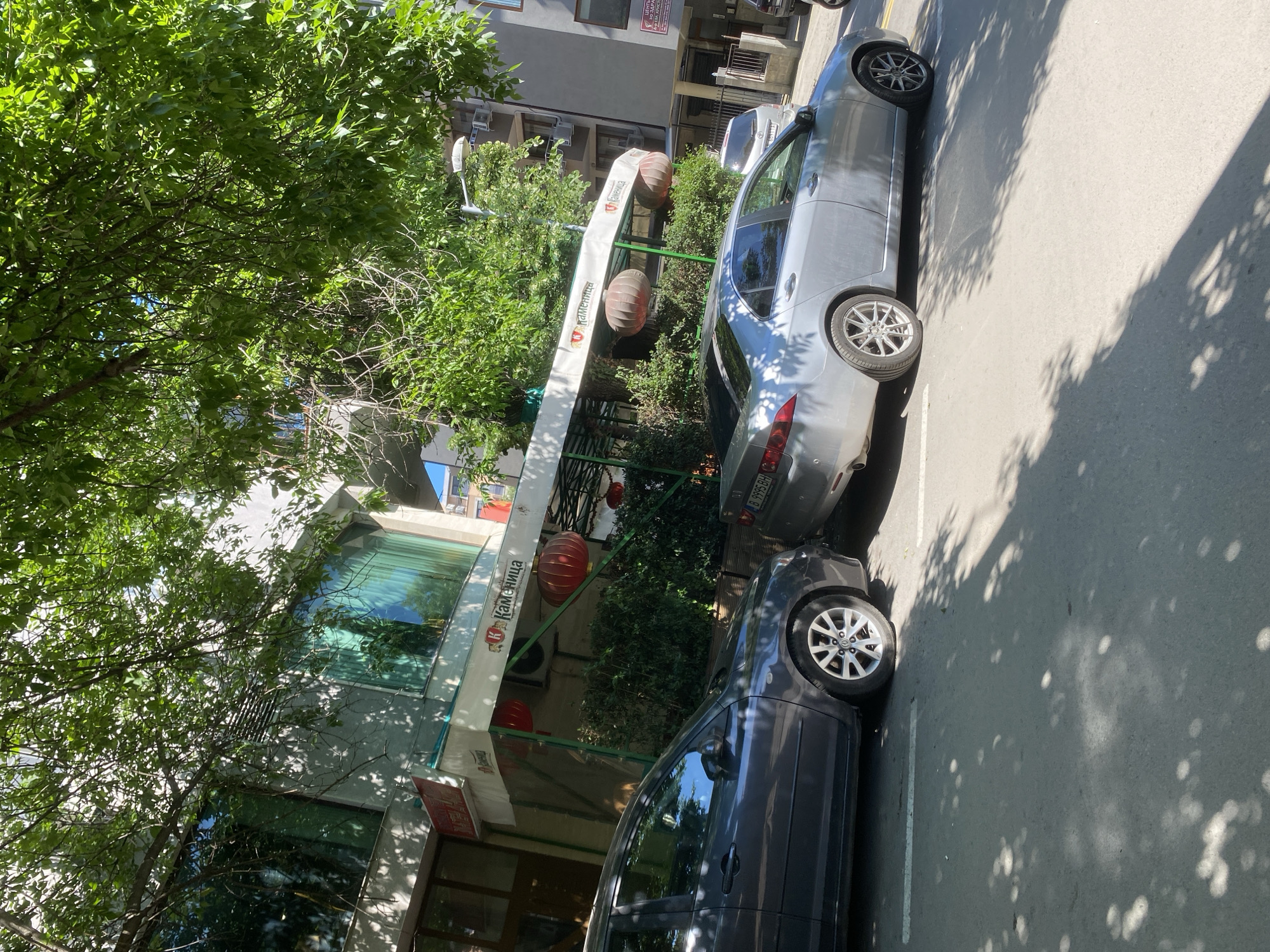 In the city of Varna, Bulgaria, on a sunny day, a gray sedan is parked on the street in front of a building. The car is positioned on the left side of the image, partially obscuring the lower portion of the building's facade. The building features a white awning with the logo "Каменица" and red decorative lanterns hanging from it. To the right of the gray sedan, a silver sedan is also parked on the street. The street is paved and marked with white lines. Lush green trees with dappled sunlight falling on the pavement are visible in the foreground and overhead. The background shows other buildings and some greenery.