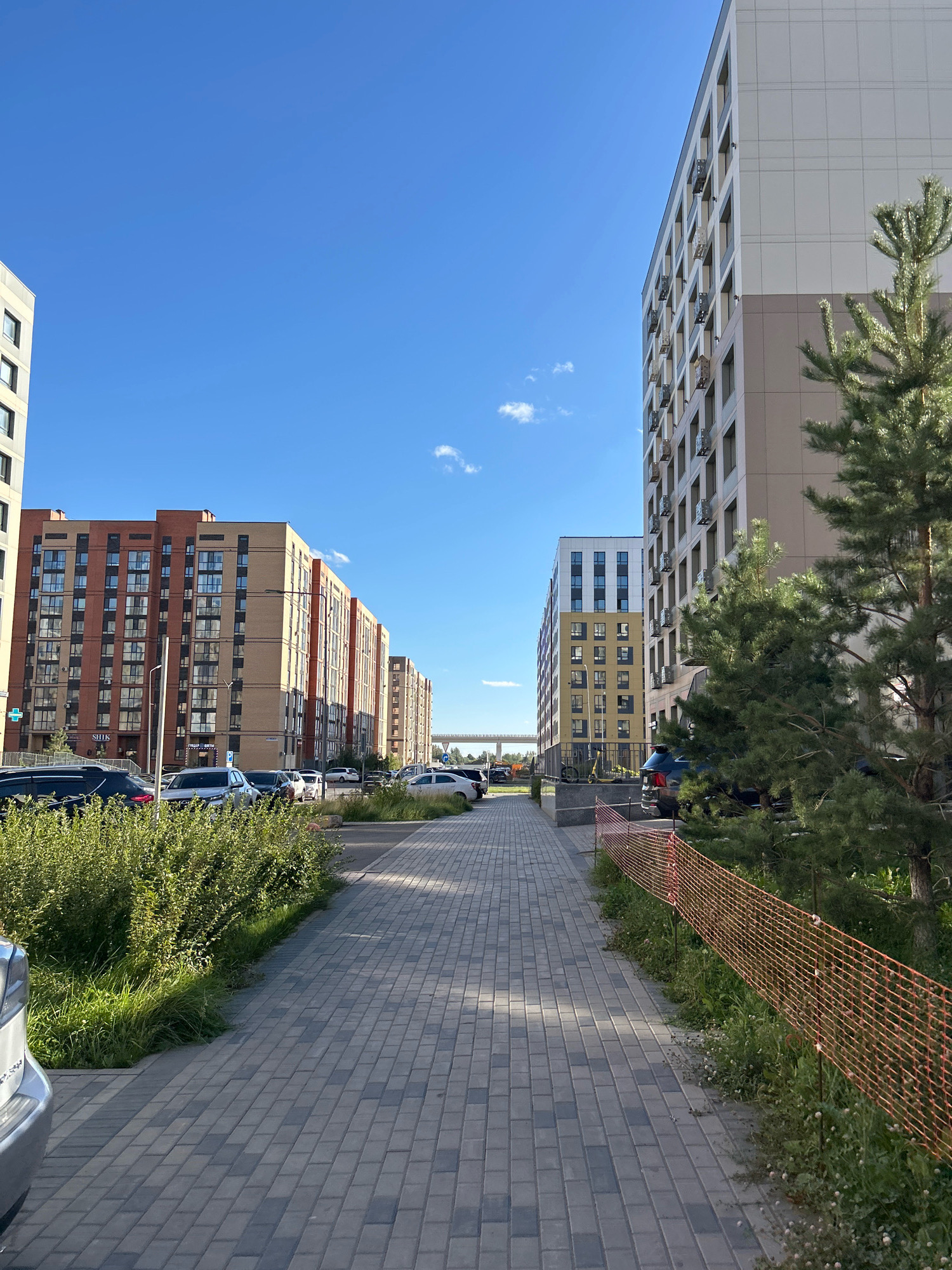 The image shows a street scene in Astana, Kazakhstan, with modern apartment buildings lining both sides of a paved walkway. On the left, buildings with red and beige facades are visible, with cars parked in front of them. Green bushes and grass line the walkway. On the right, a tall, light-colored apartment building is prominent, with a pine tree in the foreground. A temporary orange fence is also visible on the right side, along with more greenery. The sky is clear and blue, with a few scattered clouds. The scene appears to be a residential area, possibly a newly developed one, given the contemporary architecture of the buildings.