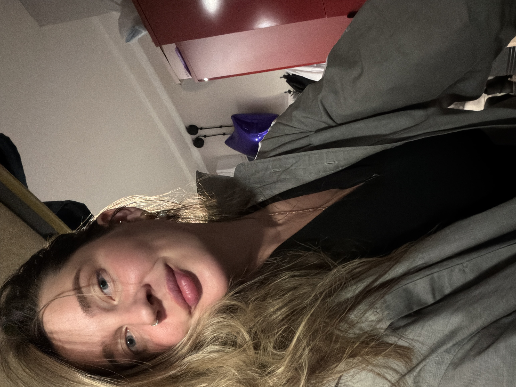 The image presents a close-up, overhead portrait of a young person, likely female, situated in an indoor setting. The subject's head is angled, looking directly towards the camera, suggesting she is lying down or leaning back. She has blonde hair, with visible darker roots, cascading around her face.
Her light-colored eyes are open, and she has a silver-toned hoop nose ring on her left nostril. Her lips are slightly parted, and a delicate gold-colored chain necklace is visible around her neck. She wears a dark-colored top underneath a light grey or olive-green collared jacket, which appears unbuttoned. One ear features a small pearl-like earring, and the other a simple hoop.
The background, partially visible, indicates an interior room. In the upper right section of the frame, a red cabinet or shelf is seen, topped with a light blue or white object. Below this, a dark blue or purple object, possibly a bag or container, rests on a surface, and two dark, circular wall-mounted fixtures are visible against a light-colored wall.
The lighting appears artificial, casting shadows and highlighting features, suggesting the photograph was taken in the evening or a dimly lit space. The emotion conveyed is neutral to slightly contemplative or inviting. No specific activities beyond posing for the photograph are evident. There are no explicit location cues within the image itself, nor is any text visible. The provided context indicates the location as Moscow, Russia.