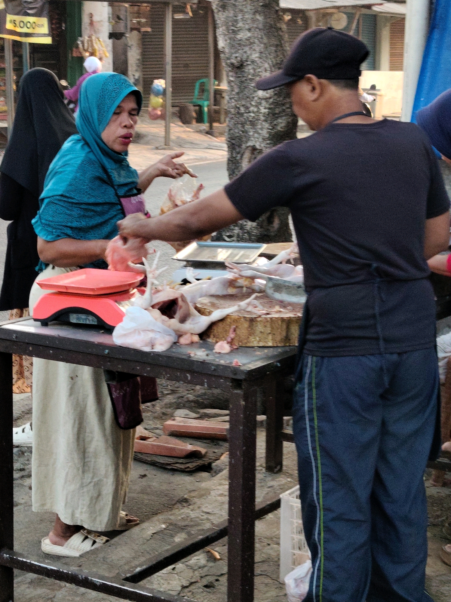 The image depicts an outdoor market scene in Purwakarta, Indonesia. In the foreground, a woman wearing a teal hijab and a long cream-colored skirt is standing behind a red weighing scale. She is holding a plastic bag and appears to be interacting with a man standing to her right. The man, who is wearing a black t-shirt and dark blue track pants with green and white stripes, has a black baseball cap on. He is facing away from the camera and appears to be involved in preparing raw chickens laid out on a wooden cutting board on the table in front of them. Other raw chickens, white plastic bags, and cutting tools are also visible on the table. In the background, a woman in a black hijab is partially visible to the left. The environment suggests a street market with various stalls and trees. The ground is paved, and there are some wooden planks and crates scattered around. A yellow sign with "5.000" is visible in the upper left corner of the image, indicating prices.