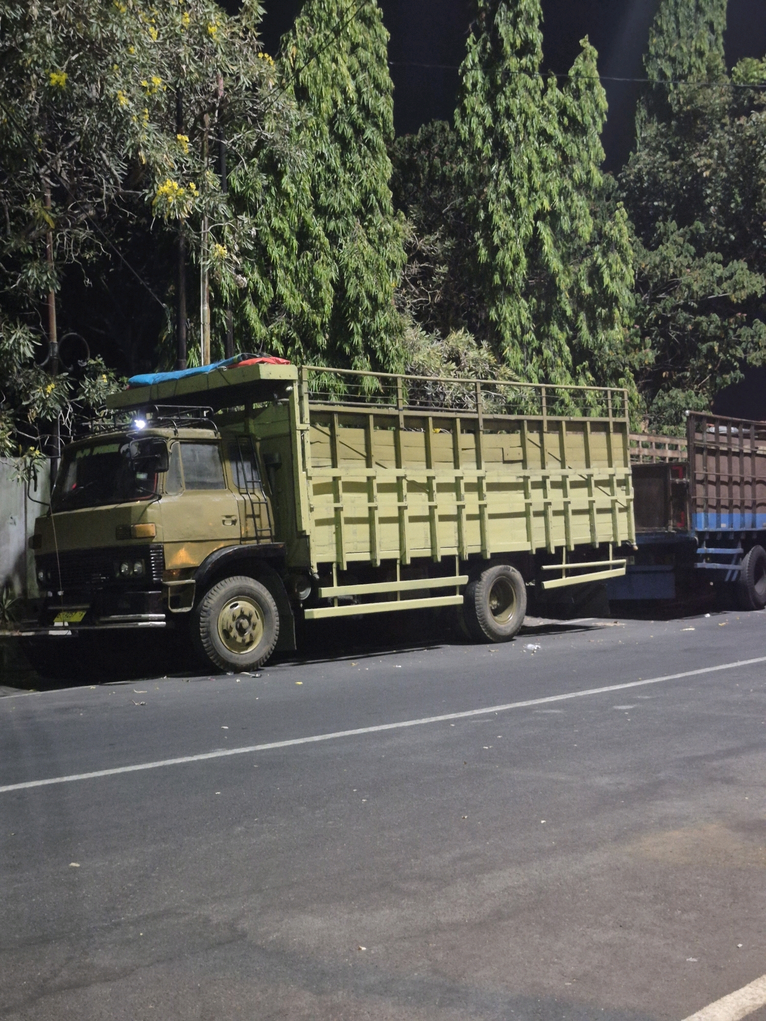 A truck and trailer combination is parked on the side of a road at night, with trees lining the background. The truck is light green and has a wooden flatbed. The trailer is dark brown and blue. There are two people visible in the image, one standing on the truck and another in the trailer. The scene appears to be in the city of Surakarta, Indonesia, as indicated by the license plate on the truck, which reads "AD 9234 AK". The overall atmosphere is dark and quiet, suggesting a late hour.