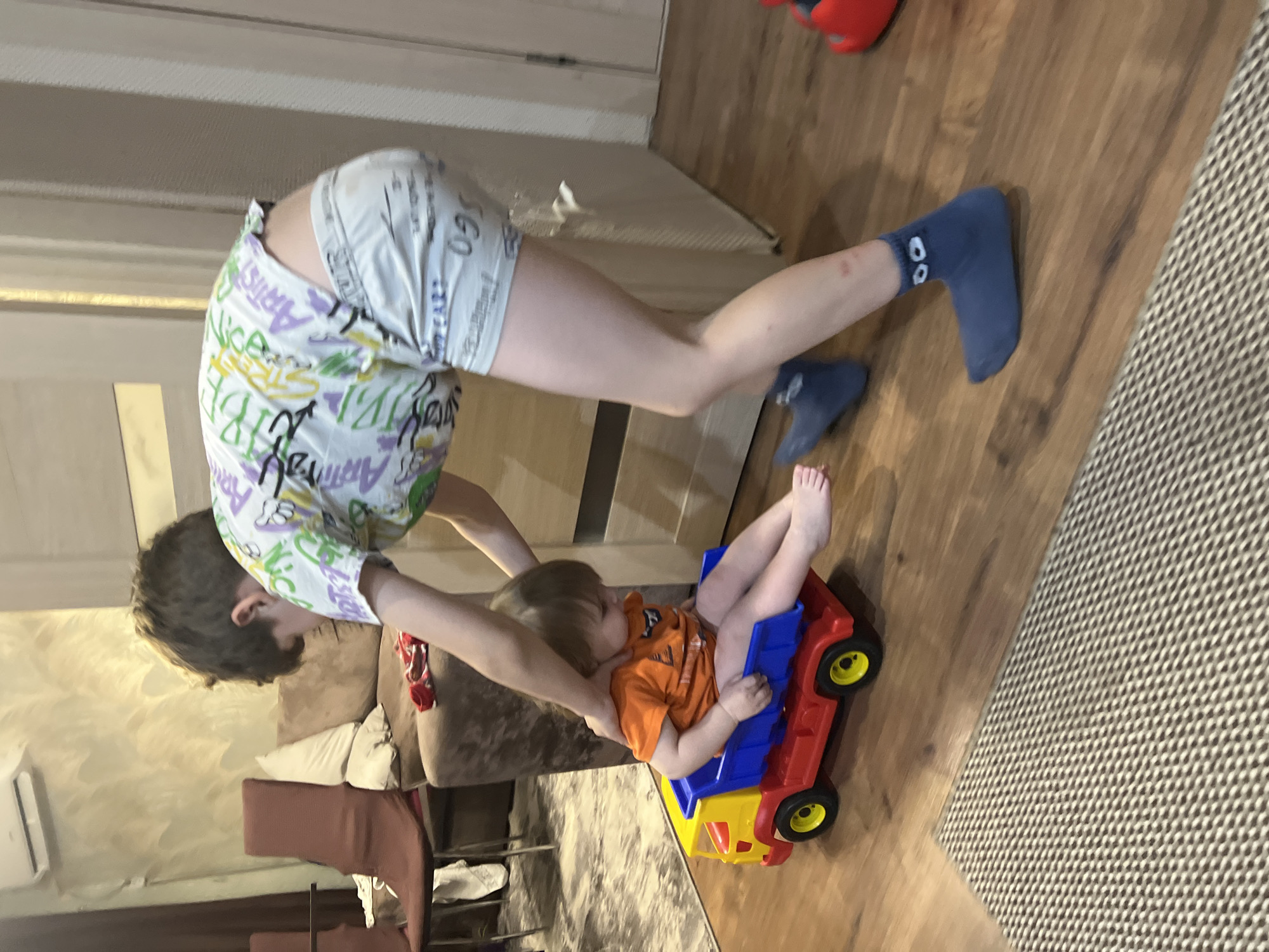 A child in shorts and socks is leaning over a smaller child sitting in a red, blue, and yellow plastic toy truck. The background shows elements of a home interior, including a couch, a rug, and a wooden floor. The image was taken indoors. The location is Noviy, Russia.
