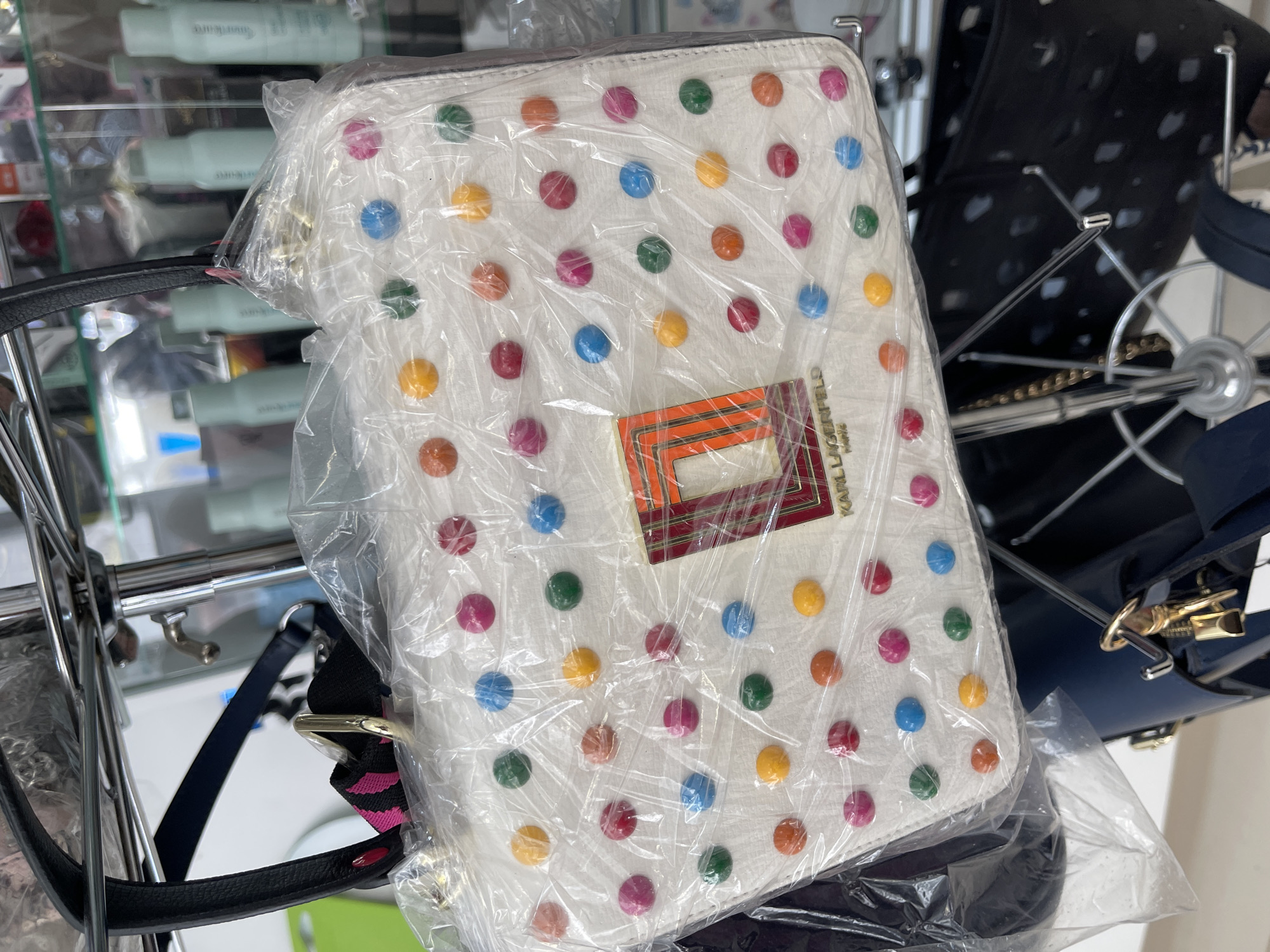 The image shows a white Karl Lagerfeld handbag covered in colorful polka dots (blue, red, pink, green, orange, yellow) inside clear plastic wrapping. The bag has a black handle and a shoulder strap with a pink and black zebra pattern detail. The background consists of a metal retail display rack holding other items, including haircare products on a glass shelf and another handbag. The environment suggests a retail setting, possibly a department store or boutique. There are no people visible. The location in Podolsk, Russia is not discernible from the image.