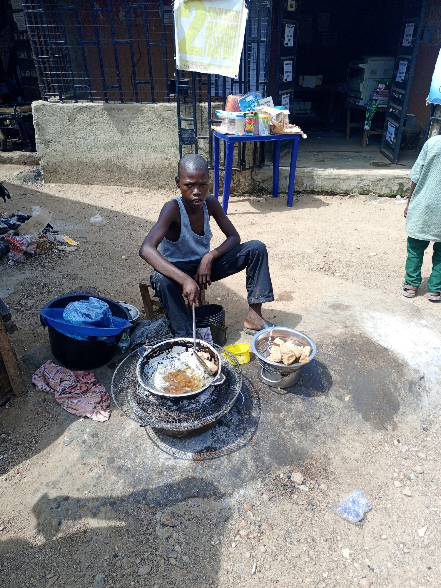 In Keffi, Nigeria, a young boy sits on a small stool, engaged in street food preparation. He wears a grey tank top and dark trousers, barefoot except for one slipper. A metal pan sits on a makeshift grill, with what appears to be oil and frying food inside. A metal colander filled with cooked food is beside the grill. A blue plastic bucket with a plastic liner sits nearby. Rubbish litters the ground. Behind him is a concrete building with barred windows and doors, indicating a possible storefront. A blue plastic table with items on it stands in front of the building. Another child stands on the right side of the frame, only their legs and part of their torso visible. The scene is outdoors on a dirt and concrete surface. It's a sunny day.